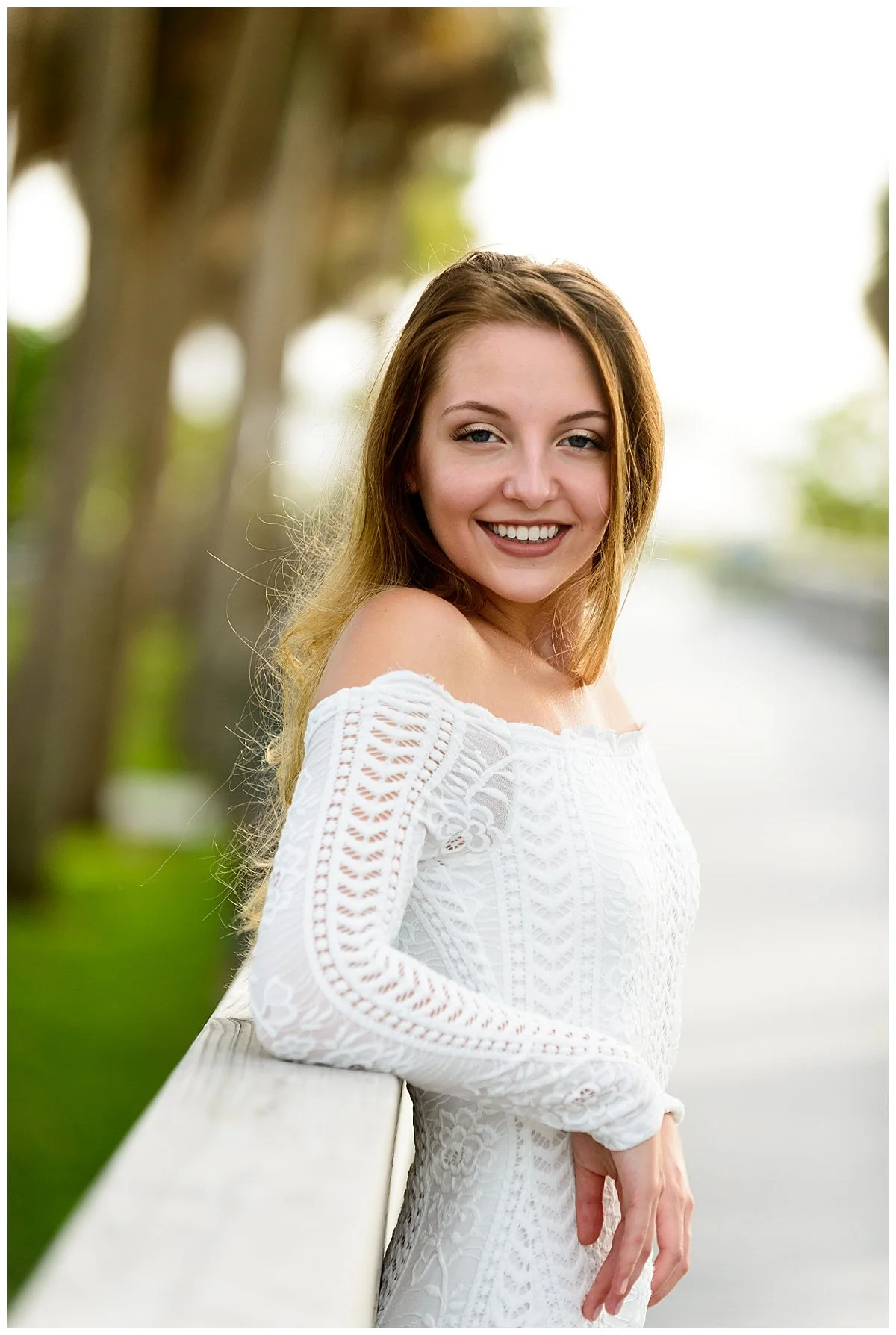 A woman with long, wavy red hair leaning on a white fence, smiling at the camera, wearing a white off-the-shoulder lace dress, outdoors with blurred trees and water in the background.