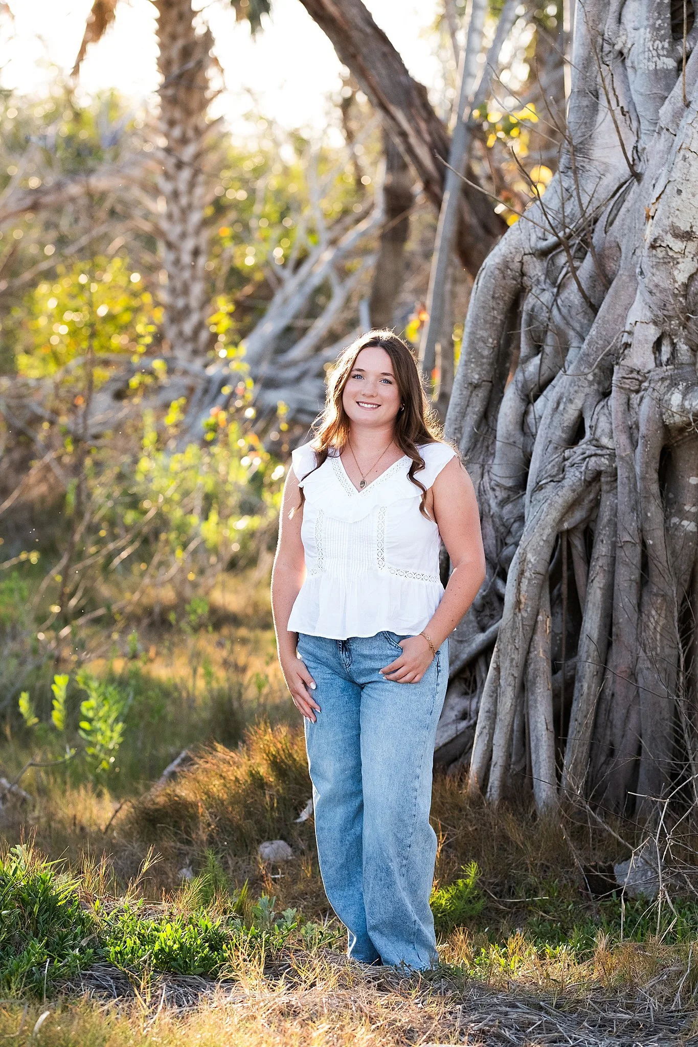 A young woman with long brown hair wearing a white sleeveless top and blue jeans, standing outdoors next to a large tree with exposed roots, smiling during golden hour.