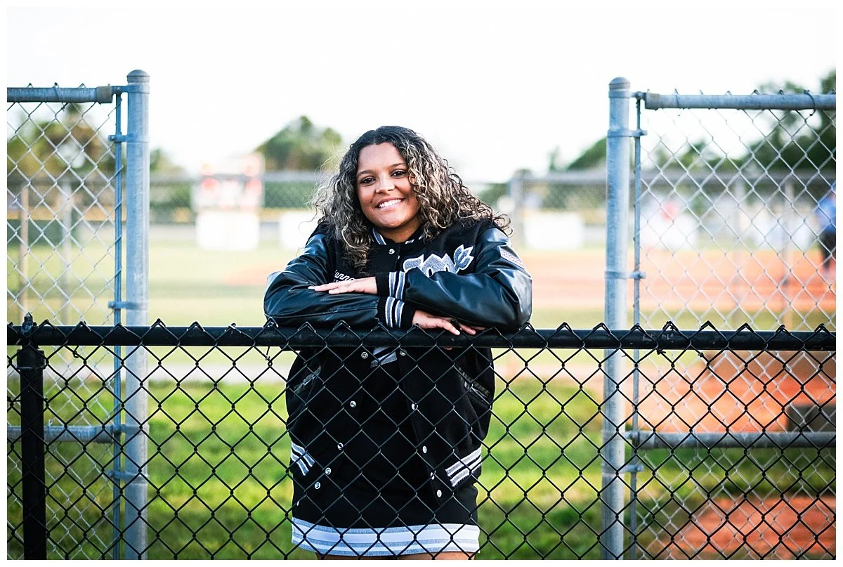 A young woman with curly hair smiling, standing behind a chain-link fence on a sports field, wearing a black jacket and a striped shirt.