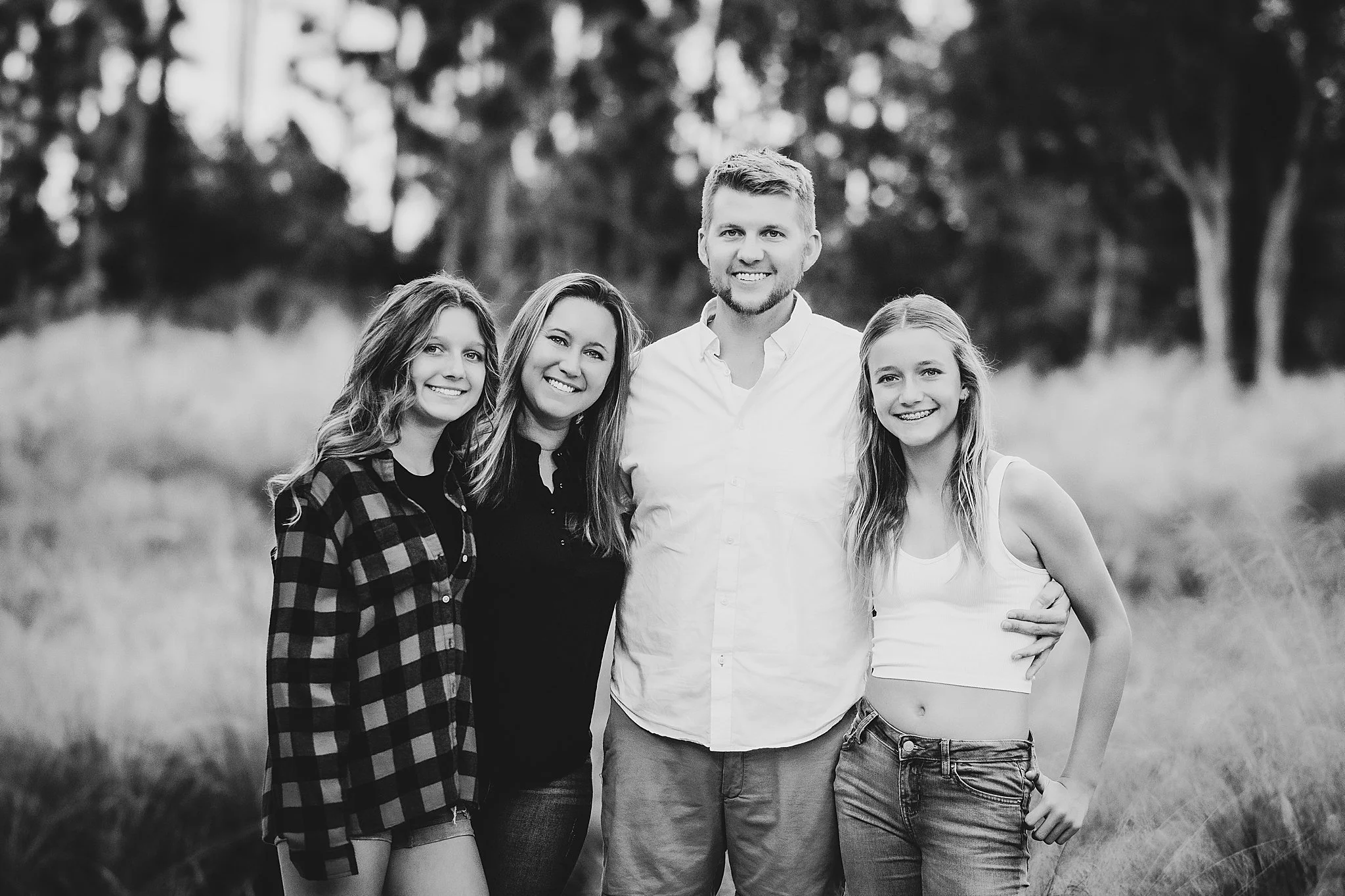 Black and white photo of a family of four outdoors, standing in a field with trees in the background. The group includes two women, a man, and a teenage girl, all smiling and close together.