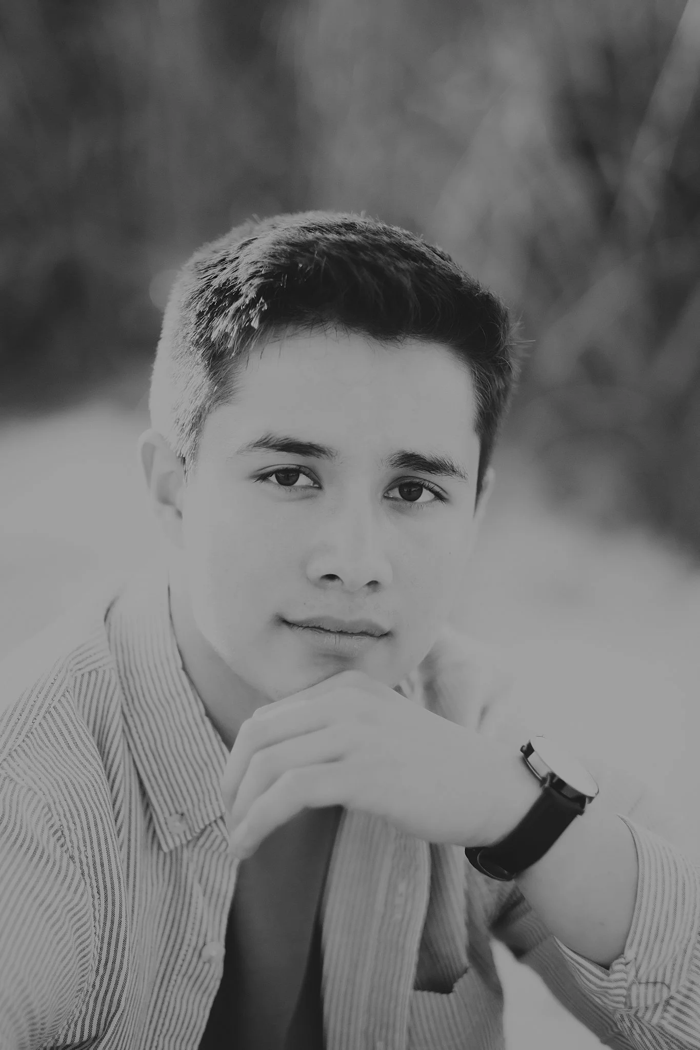 Portrait of a young man with short dark hair and light skin, wearing a striped shirt and a watch, resting his chin on his hand outdoors.