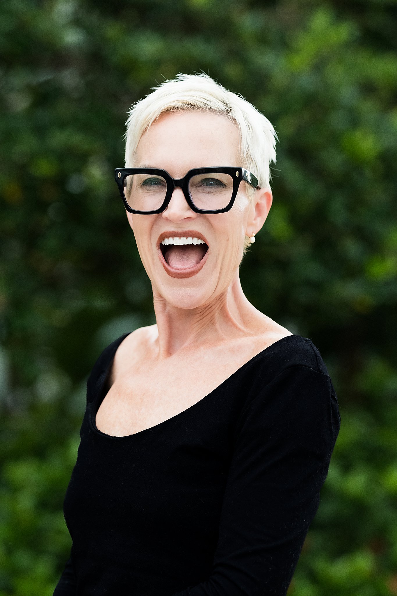 A woman with short platinum blonde hair wearing large black glasses and a black top, smiling widely outdoors with greenery in the background.