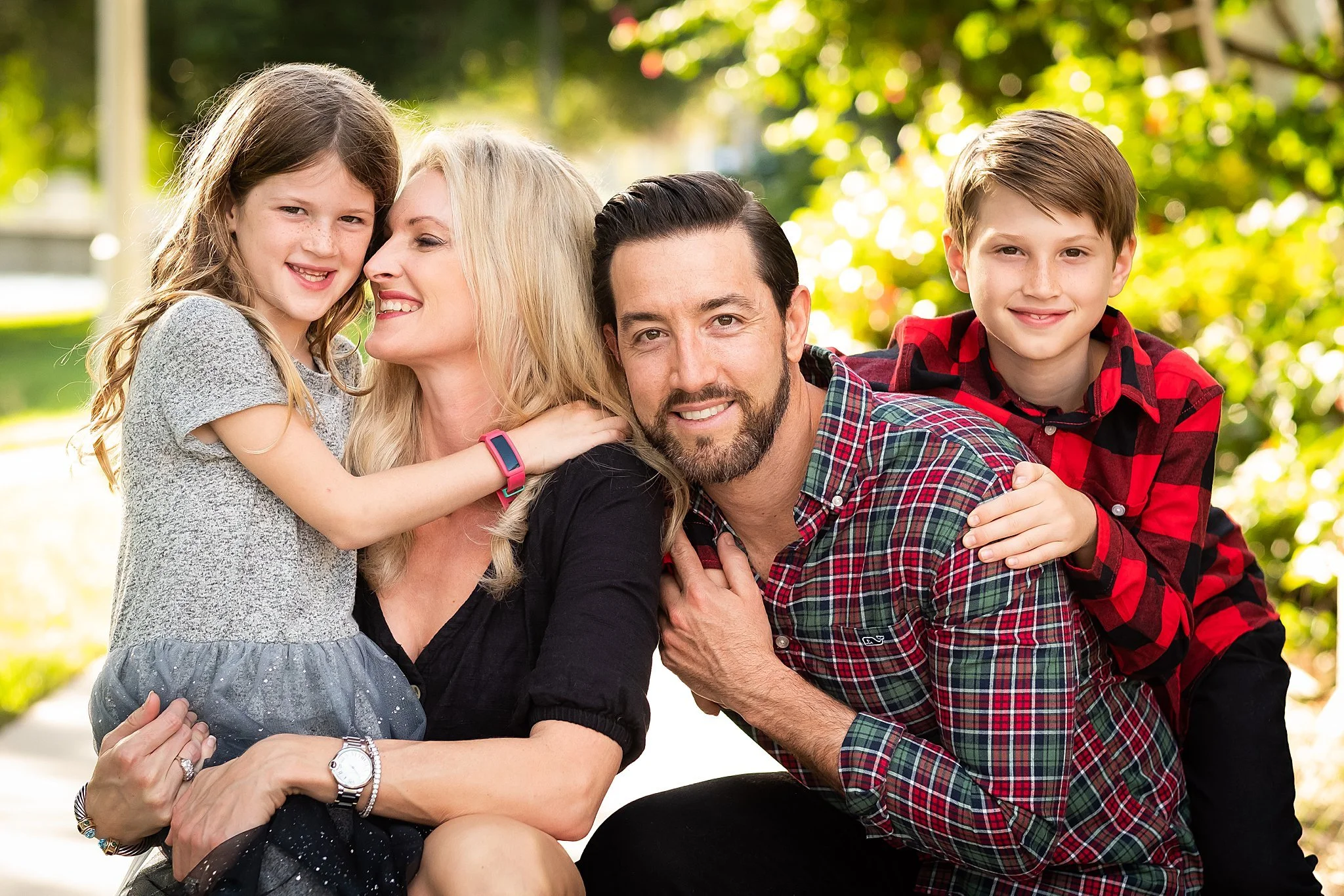 Family of four outdoors, mother with daughter and father with son, smiling and hugging each other in a park with green trees in the background.
