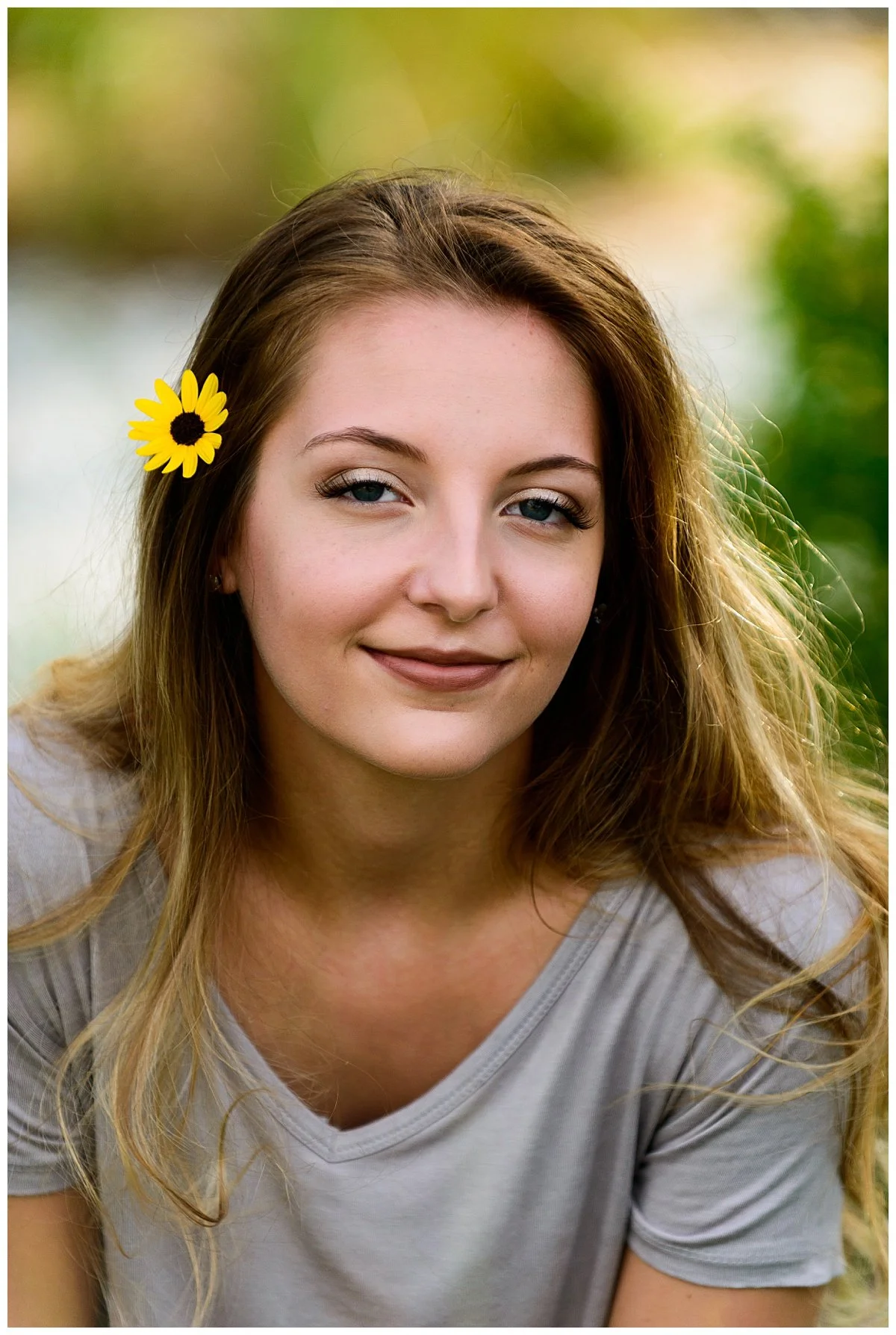 A young woman with long, wavy hair, a yellow flower in her hair, and a light gray shirt, outdoors with a blurred background of greenery.
