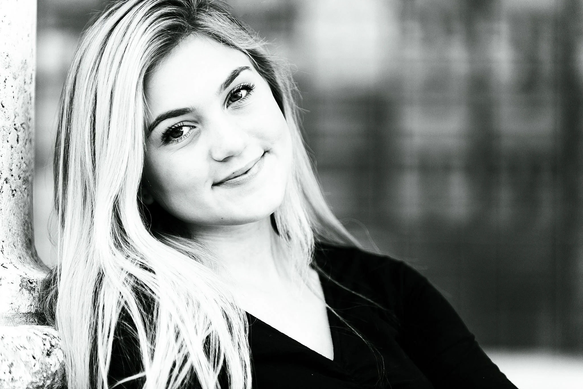 Black-and-white portrait of a young woman with long blonde hair, leaning against a brick wall, smiling softly at the camera.
