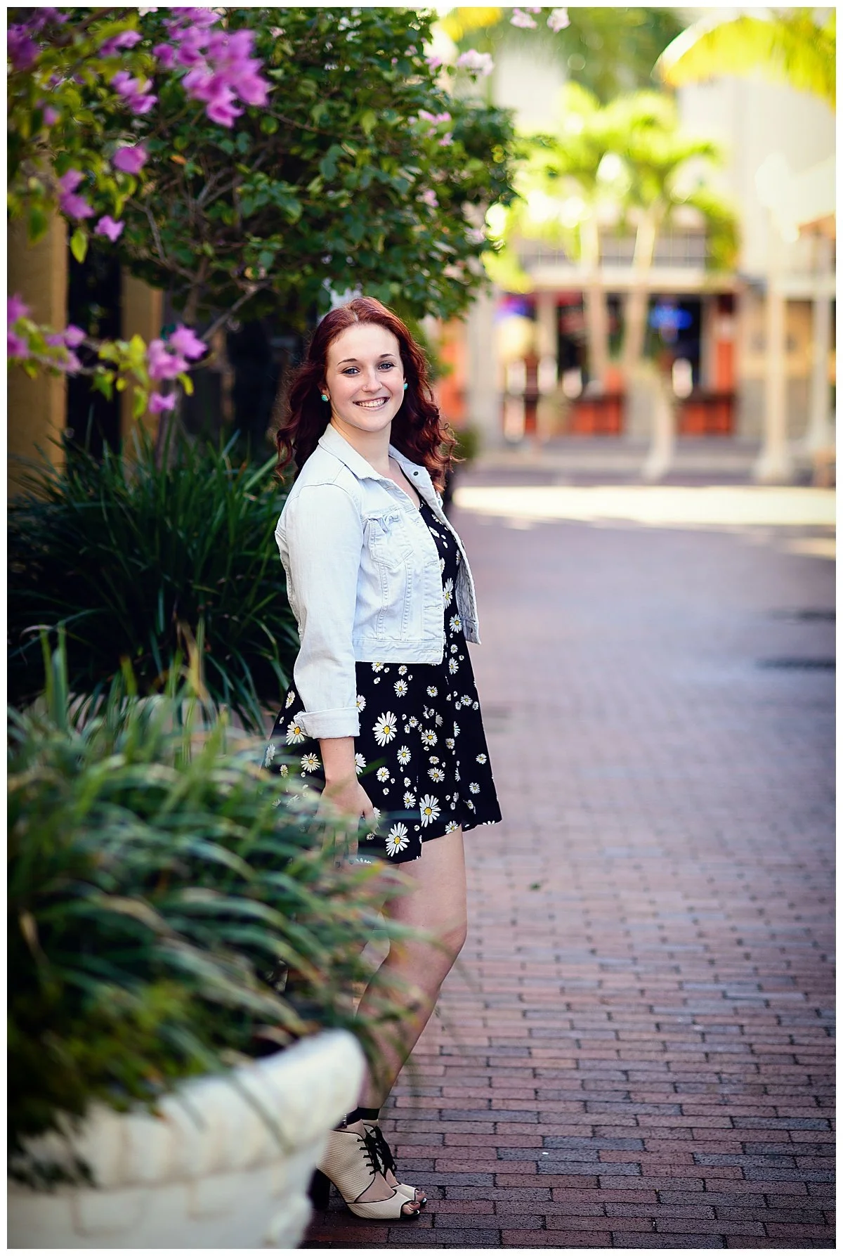 A young woman with red hair stands on a brick sidewalk, smiling at the camera. She wears a black floral dress with daisies, a light denim jacket, and beige high heels. There are green plants and a blooming pink bougainvillea behind her, with a blurre