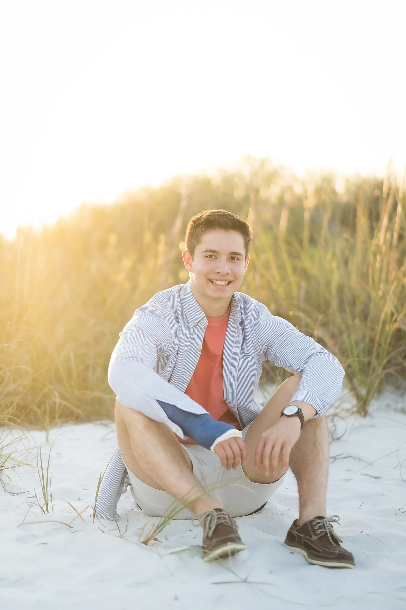 A young man sitting on the sand at the beach during sunset, smiling at the camera, wearing a striped button-down shirt, a watch, and casual shoes, with tall grass in the background.