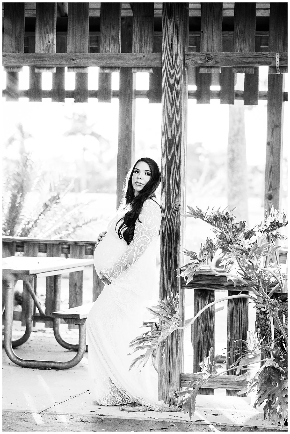 A pregnant woman dressed in a long lace dress stands outdoors under a wooden railing, holding her belly and looking at the camera. The background features blurred trees and natural scenery, captured in black and white.