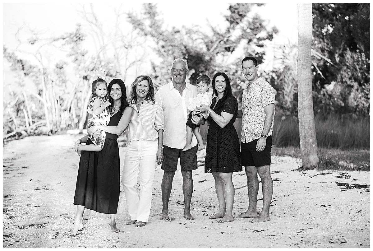 Family portrait of seven people standing on a sandy beach, smiling and posing for the photo, with trees in the background. It appears to be a multi-generational family, including children, adults, and seniors.