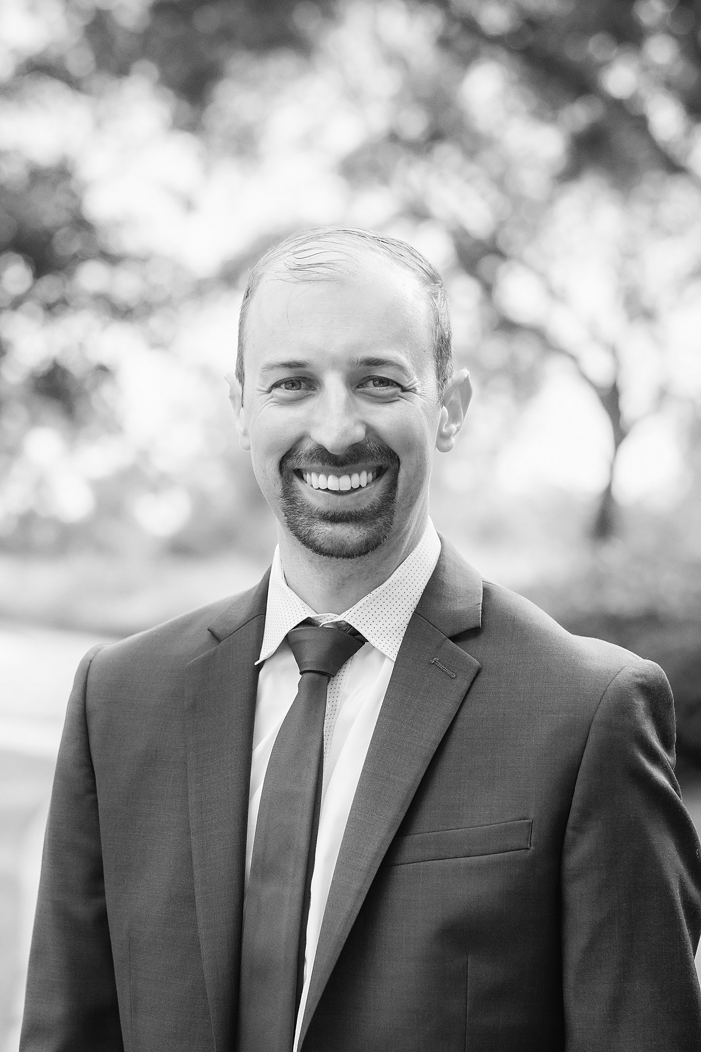 A smiling man in a suit and tie outdoors, blurred trees in the background.