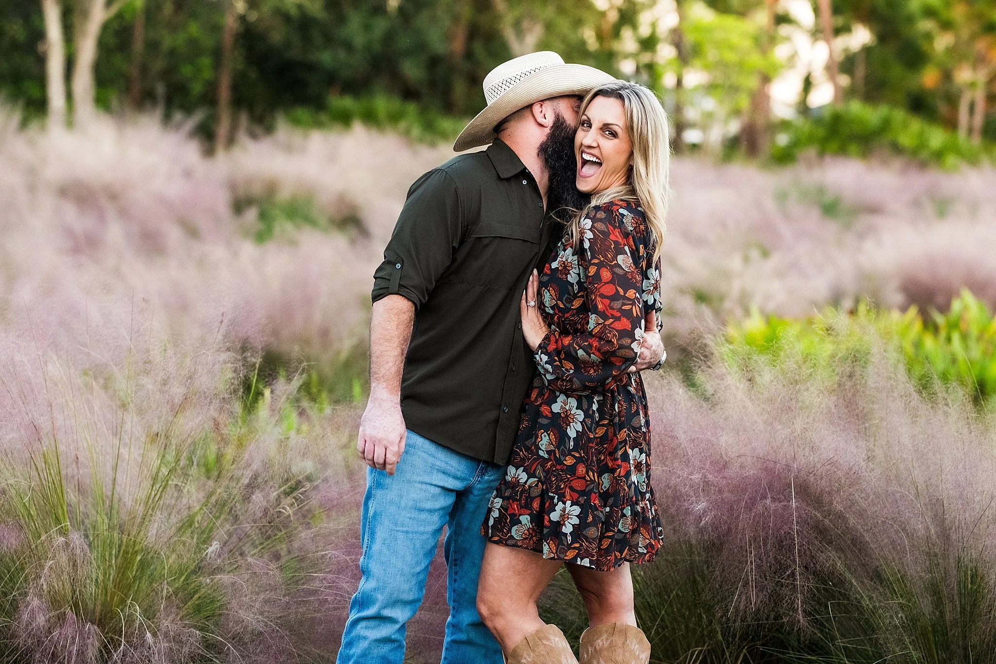 A smiling woman in a floral dress and cowboy boots posing with a bearded man in a dark shirt and hat, standing in a field of pinkish-purple grass.