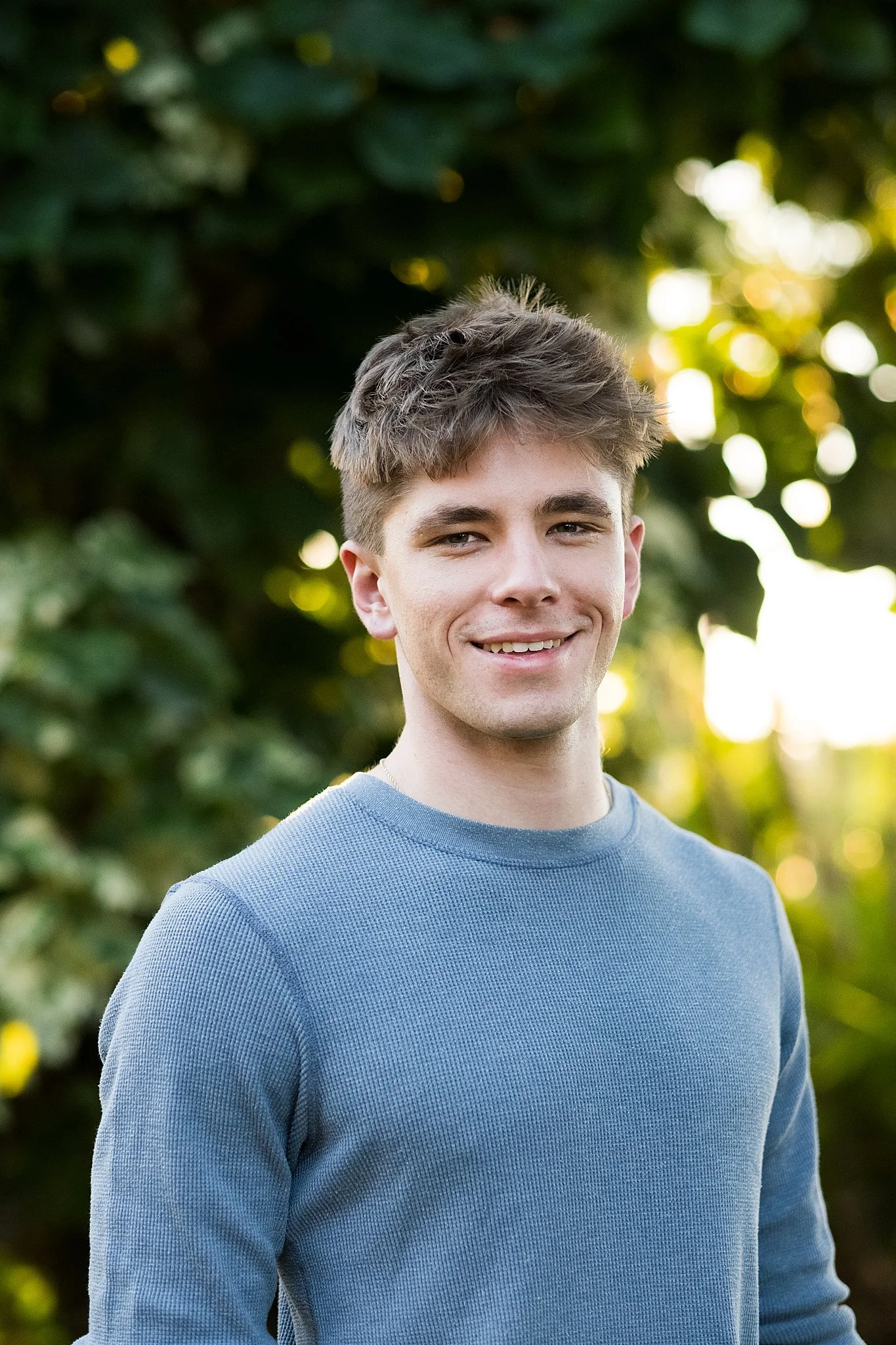 A young man with short brown hair and light skin, smiling, wearing a light blue sweater, outdoors with green foliage and sunlight in the background.