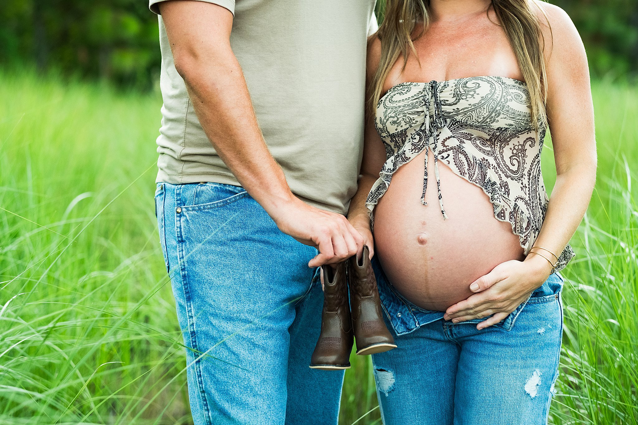 A pregnant woman standing outdoors in a grassy field, holding a pair of small brown boots, with a man standing beside her and her hand resting on her belly.