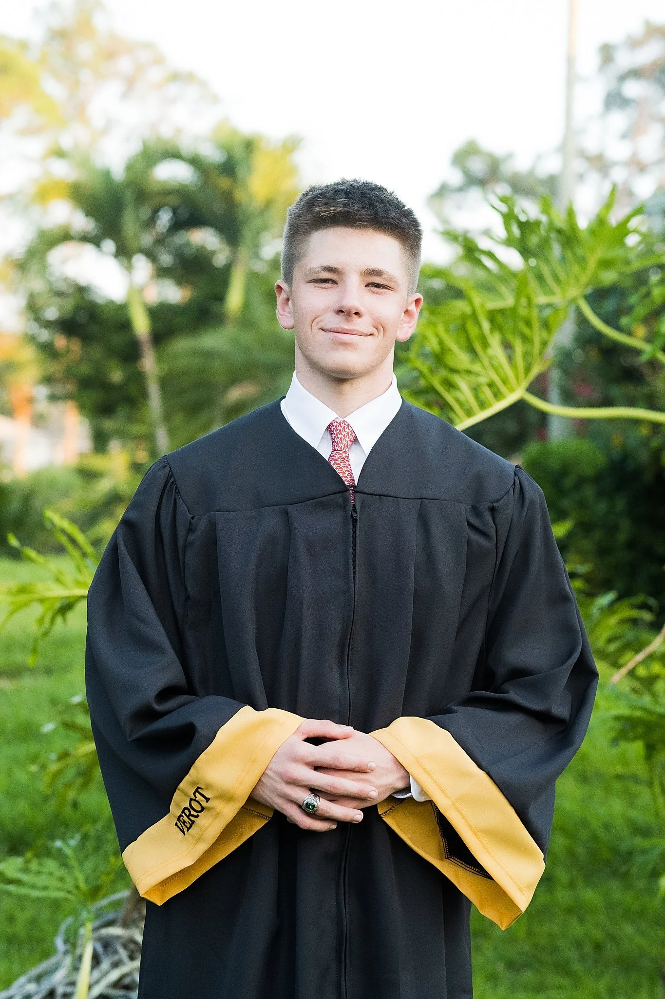 Young man in graduation gown outdoors with greenery in background.