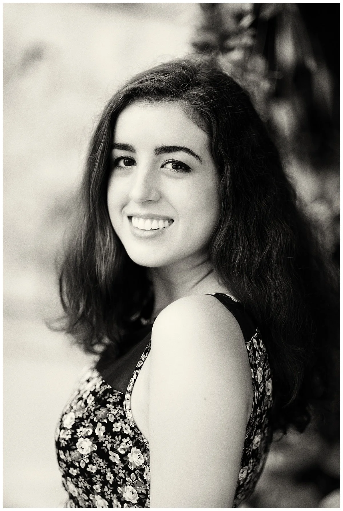 A black-and-white portrait of a young woman with long, wavy hair smiling at the camera, wearing a sleeveless floral dress.