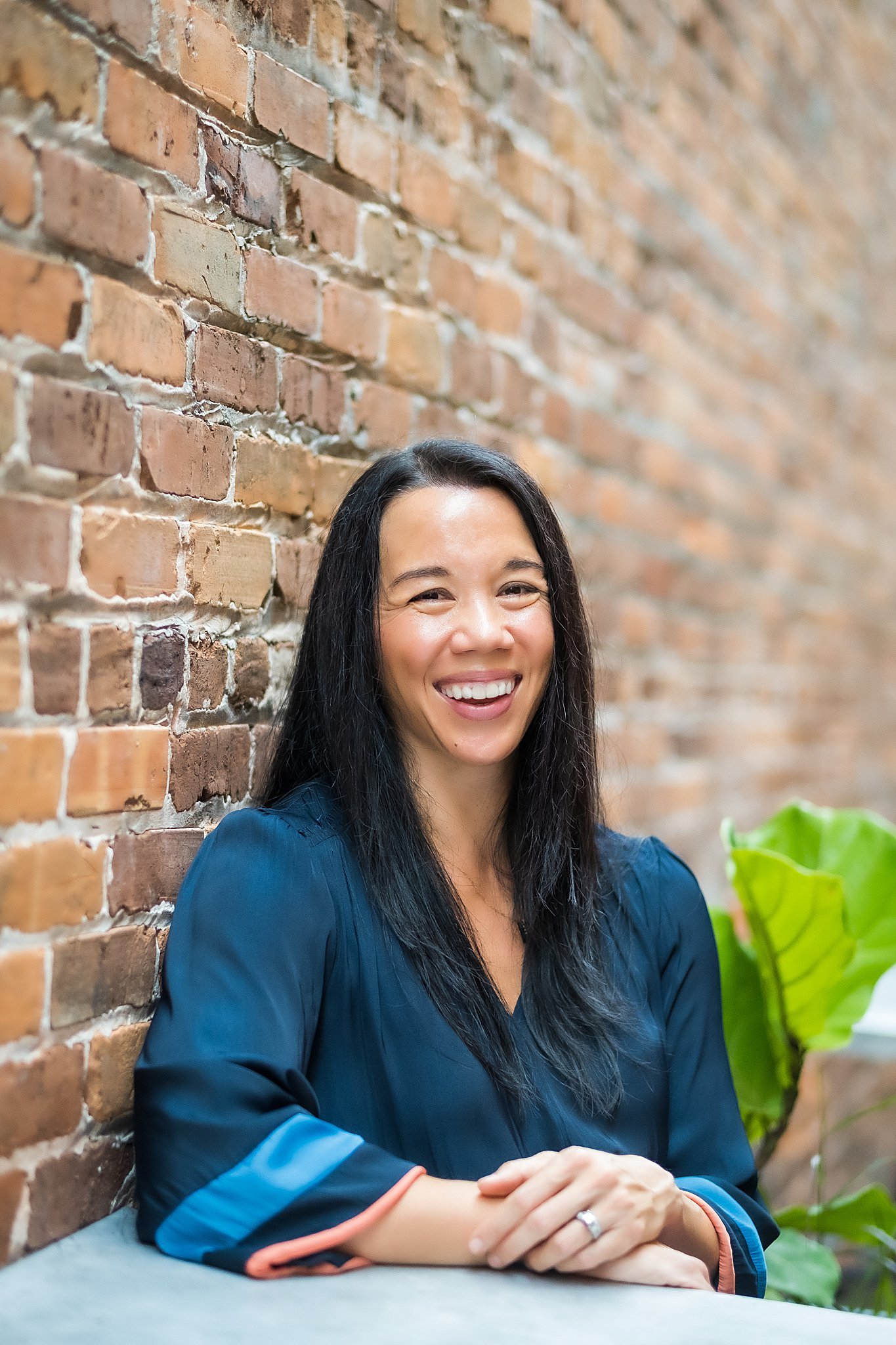 A woman with long black hair smiling and sitting in front of a brick wall, wearing a dark blue satin blouse.
