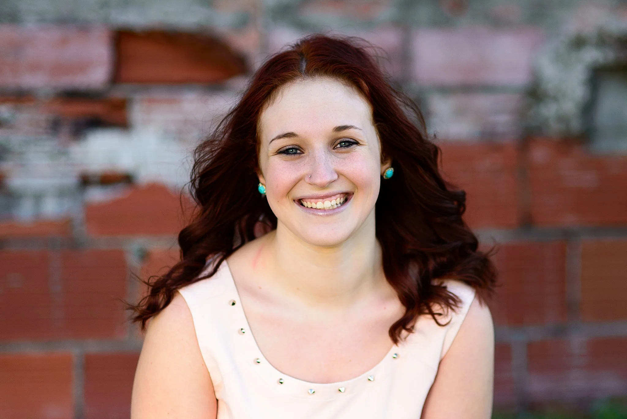 A young woman with red hair smiling outdoors, wearing a sleeveless beige top with small decorative studs, turquoise earrings, and a nose piercing, standing in front of a brick wall.