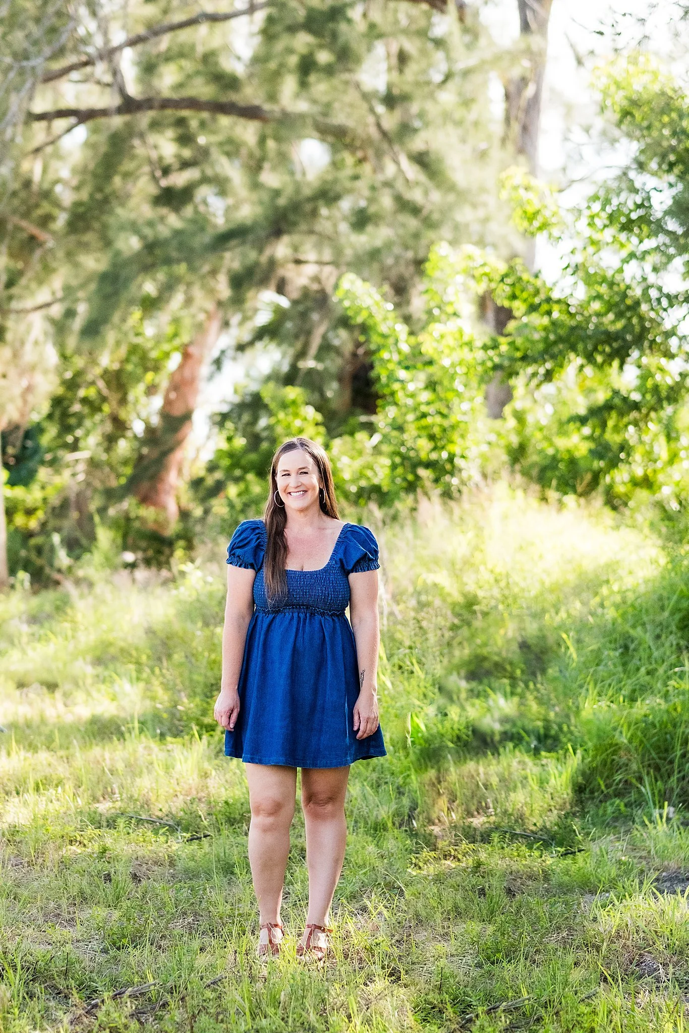A woman with long brown hair wearing a blue dress and sandals standing outdoors in a grassy area with trees and sunlight in the background.