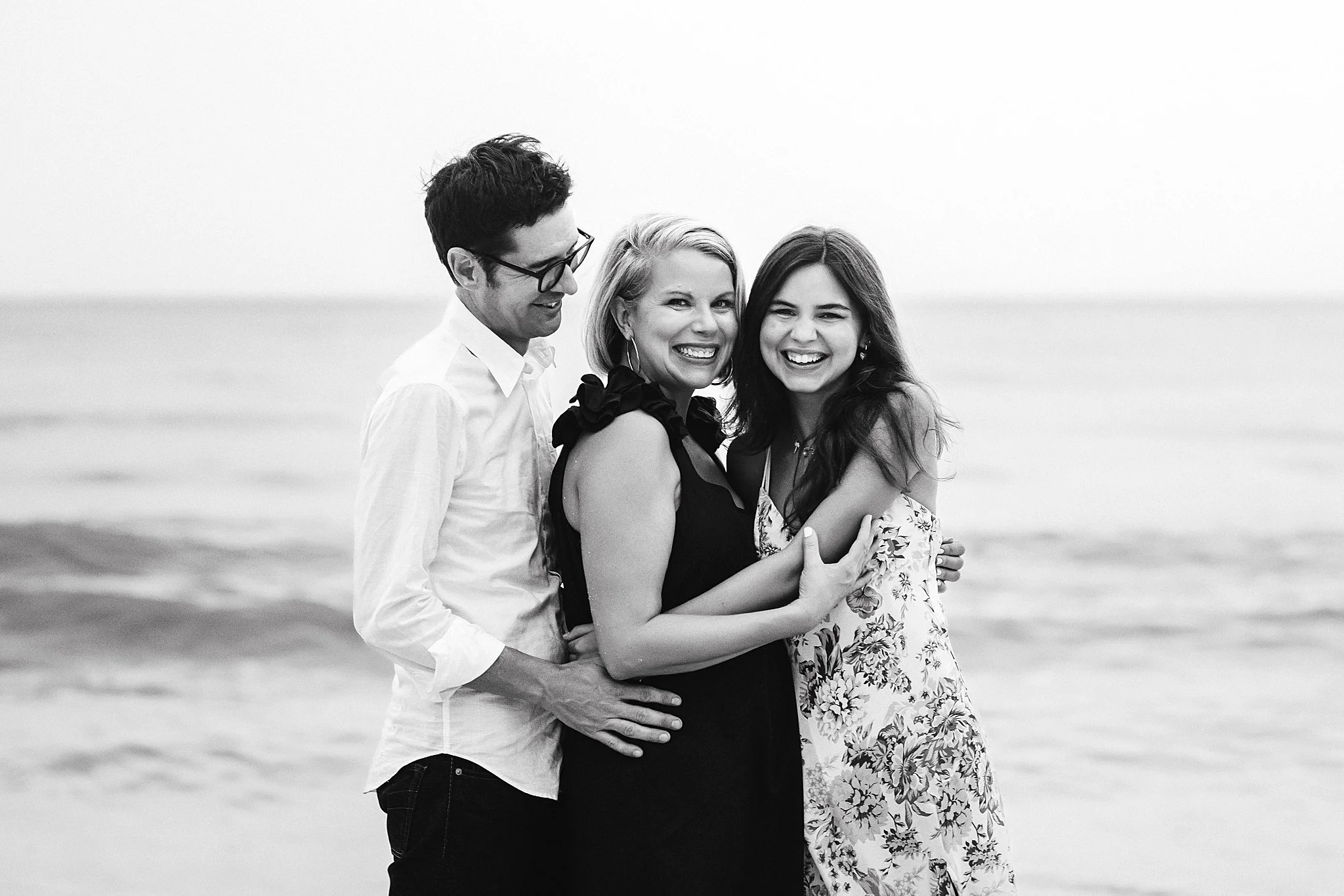 A group of three people, two women and one man, smiling and hugging on a beach. The photo is in black and white.