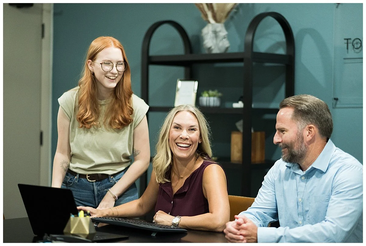 Three people in an office or meeting room, smiling and laughing. One woman is standing, and the other two are sitting at a table with a laptop and a keyboard. The background features a black shelving unit with decorative items.