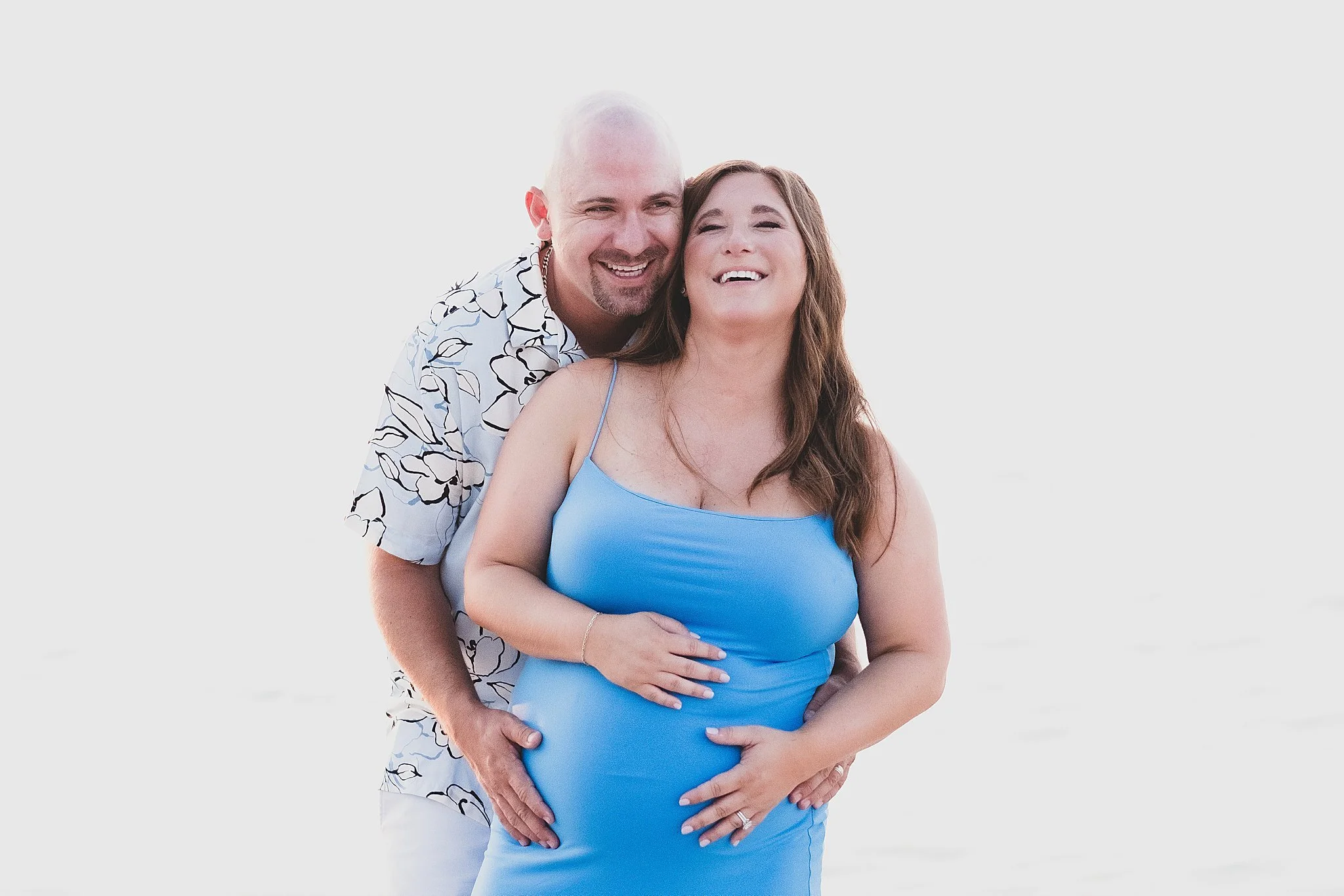 A smiling couple, a pregnant woman in a blue dress and a man in a patterned shirt, embracing on a bright beach with white sand and clear sky.