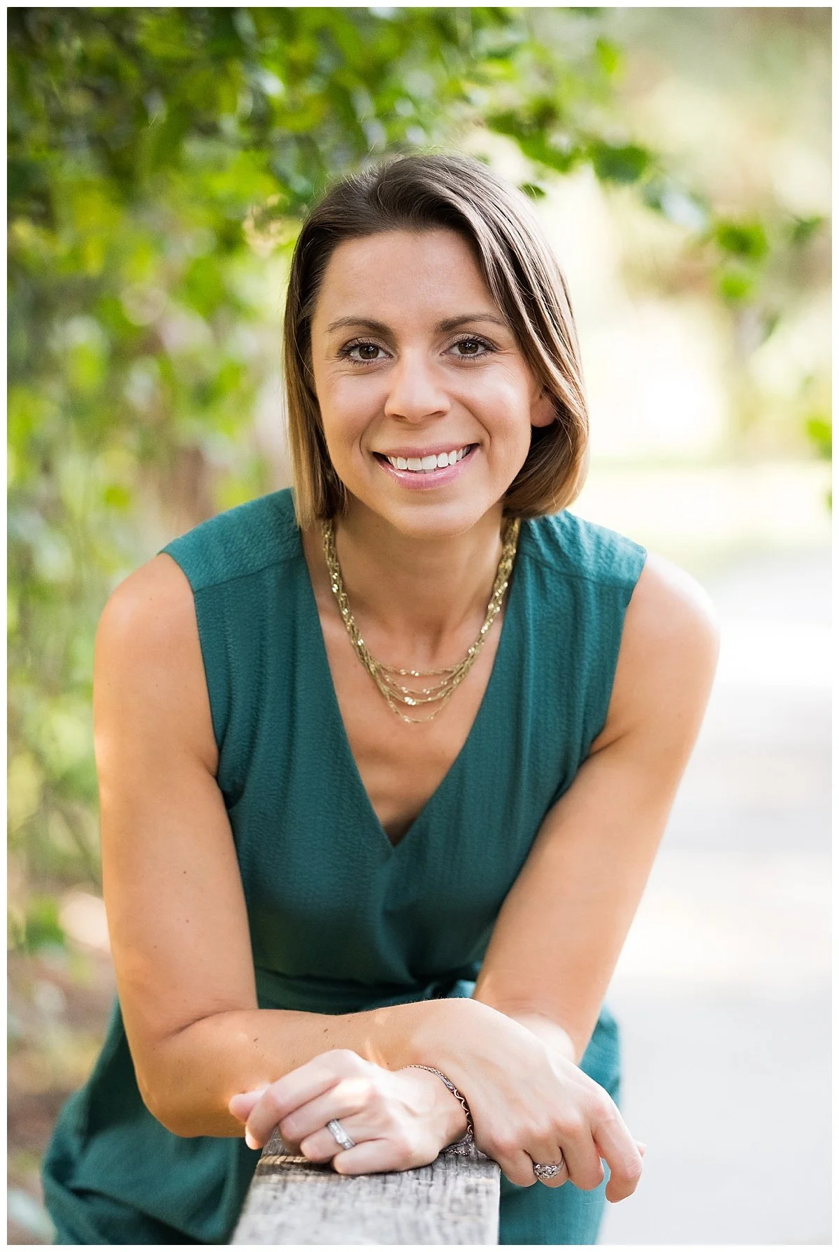 A smiling woman with short brown hair wearing a sleeveless teal top, gold necklaces, and rings, leaning on a wooden railing outdoors with green foliage in the background.