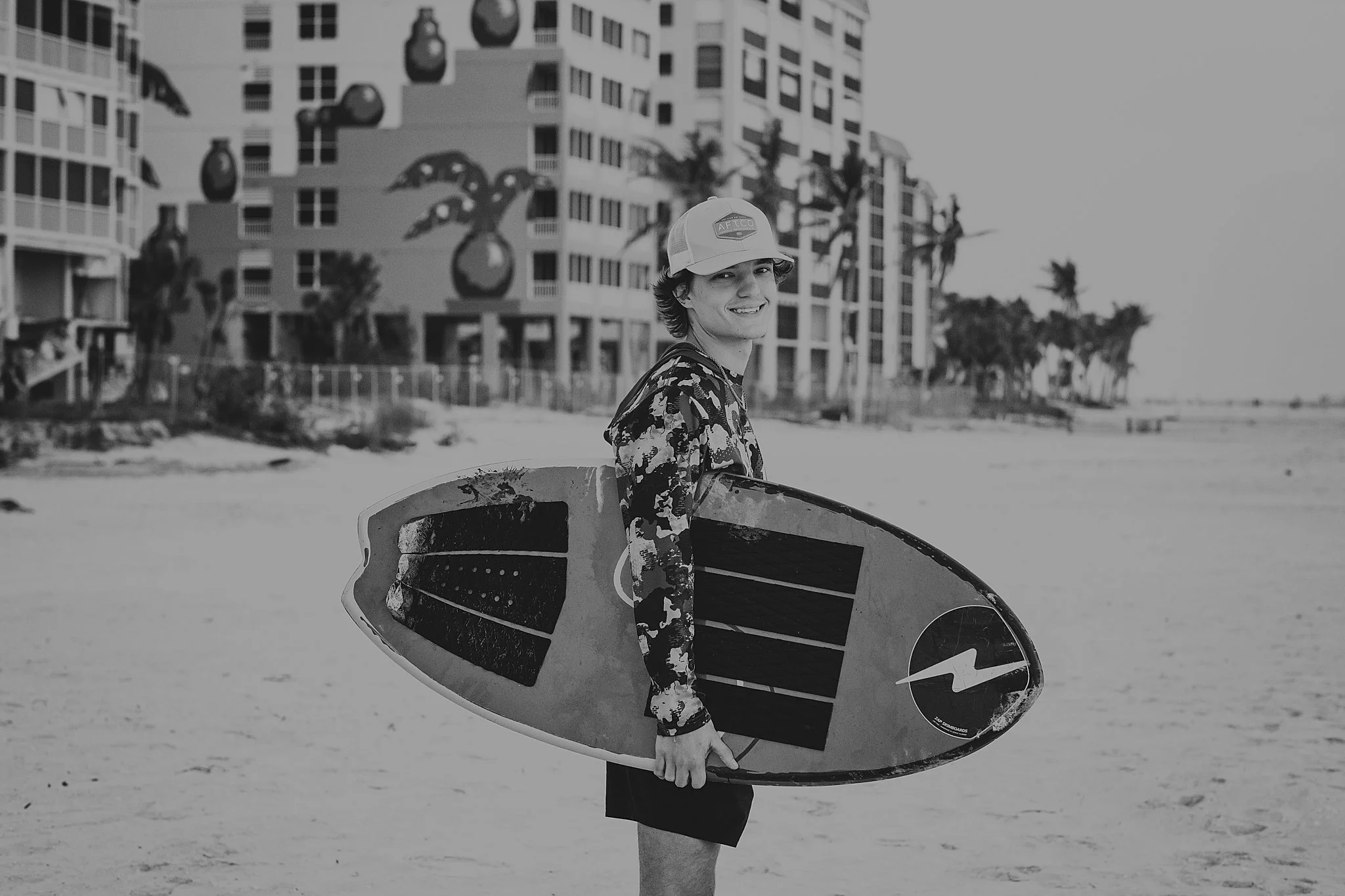 A young man holding a surfboard on a beach with buildings and palm trees in the background.