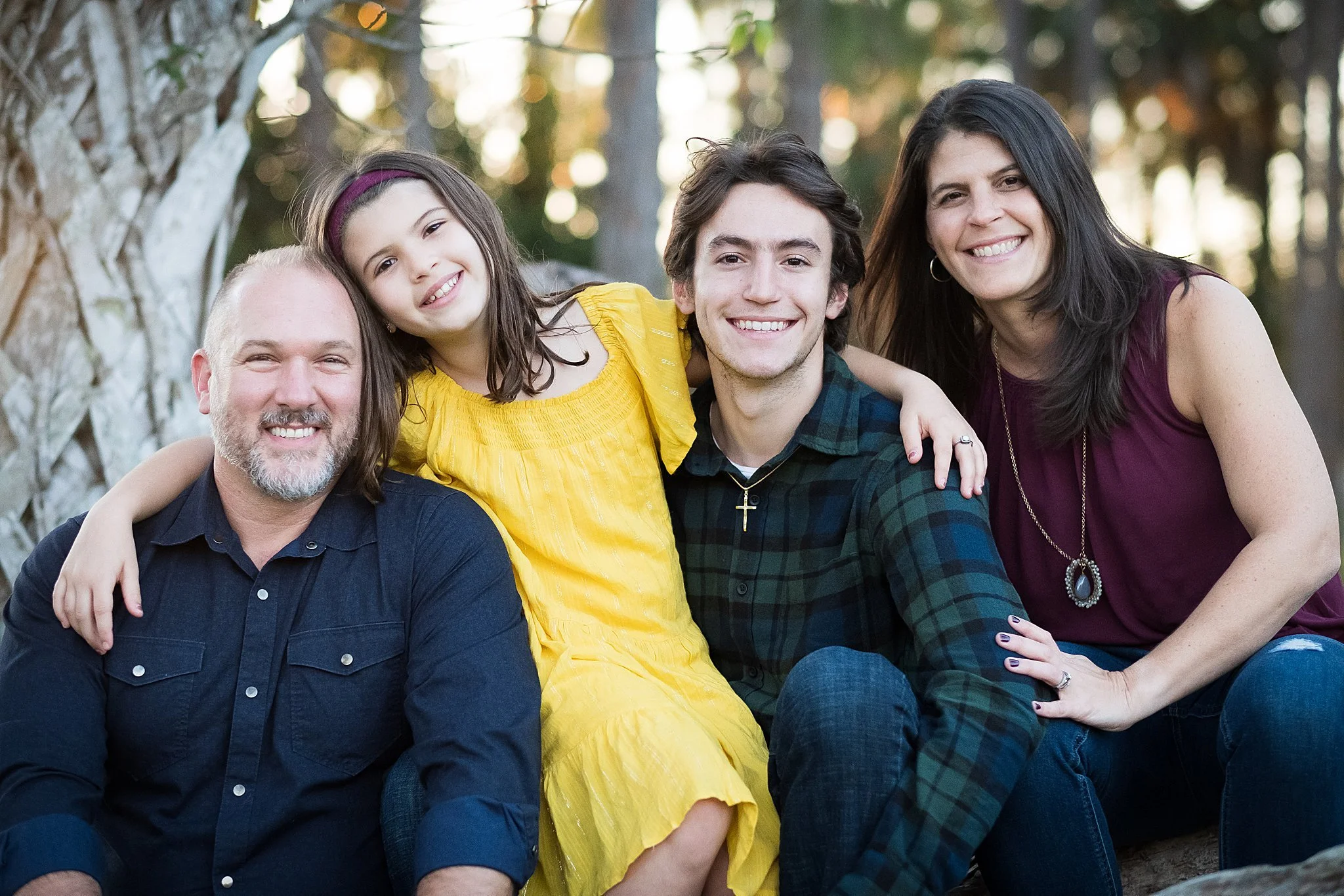 A smiling family of four sitting outdoors in front of trees, with a man, a woman, a teenage boy, and a young girl, all dressed casually.