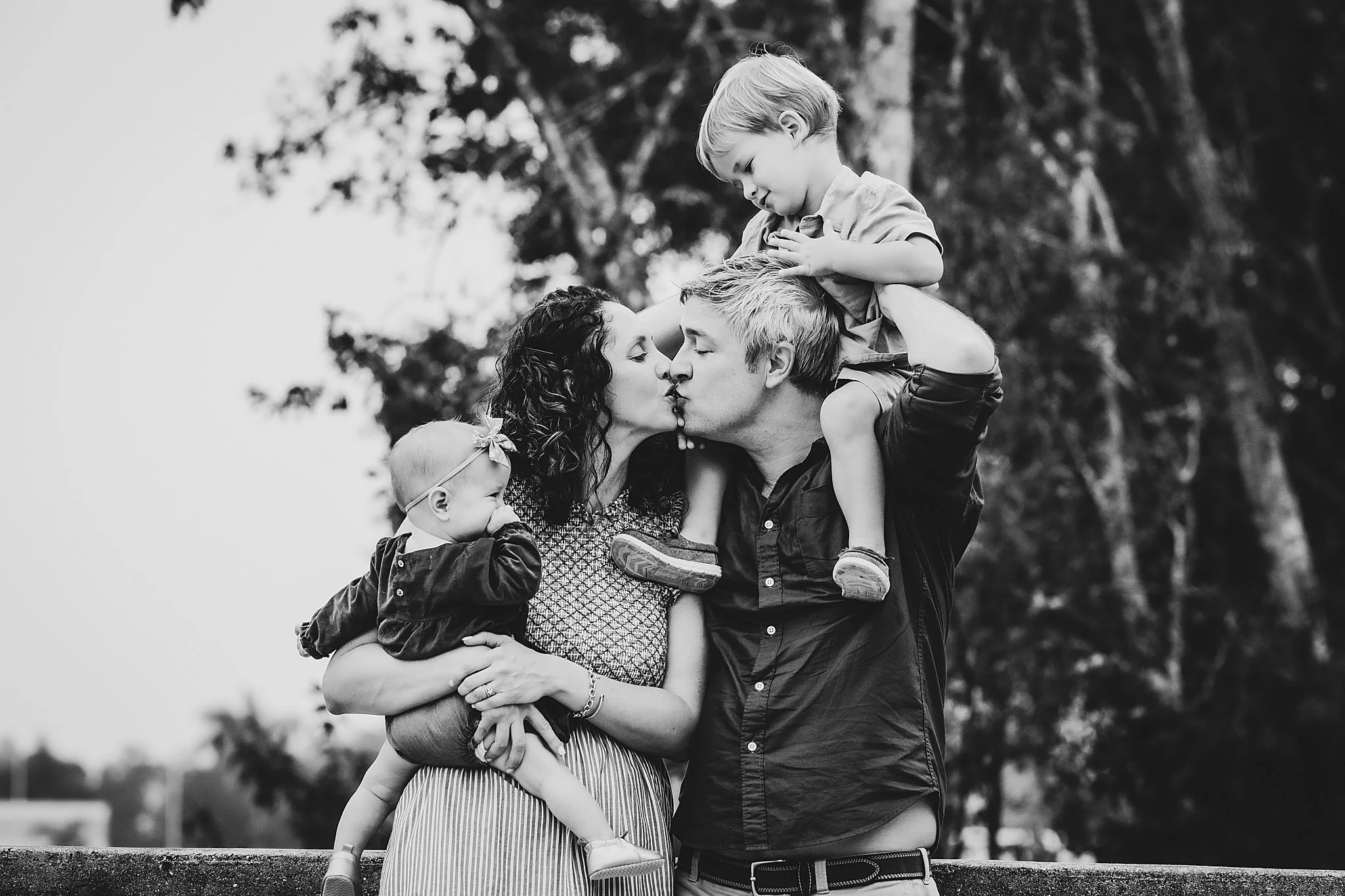 A family of four sharing a kiss outdoors, with a mother, father, and two young children, in black and white.