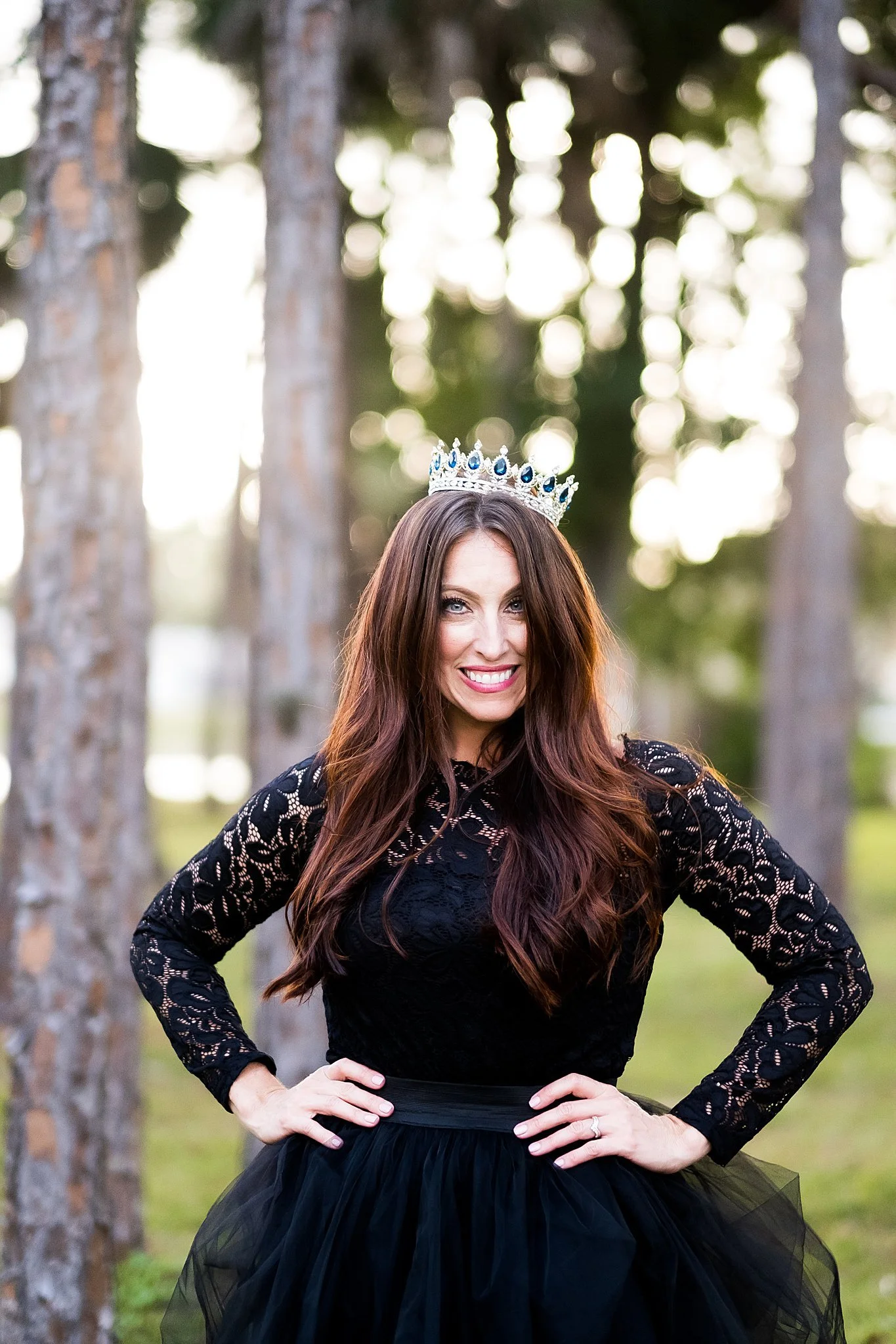 A woman in a black lace dress and black tulle skirt wearing a jeweled crown, standing outdoors among trees, smiling with hands on her hips.