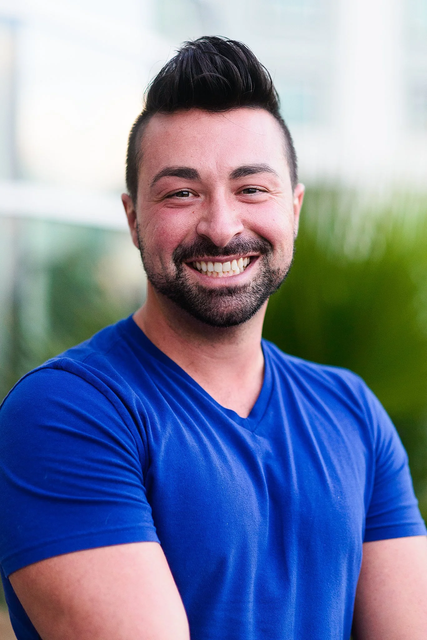 A smiling man with dark hair and a beard, wearing a blue T-shirt, standing outdoors with blurred greenery in the background.