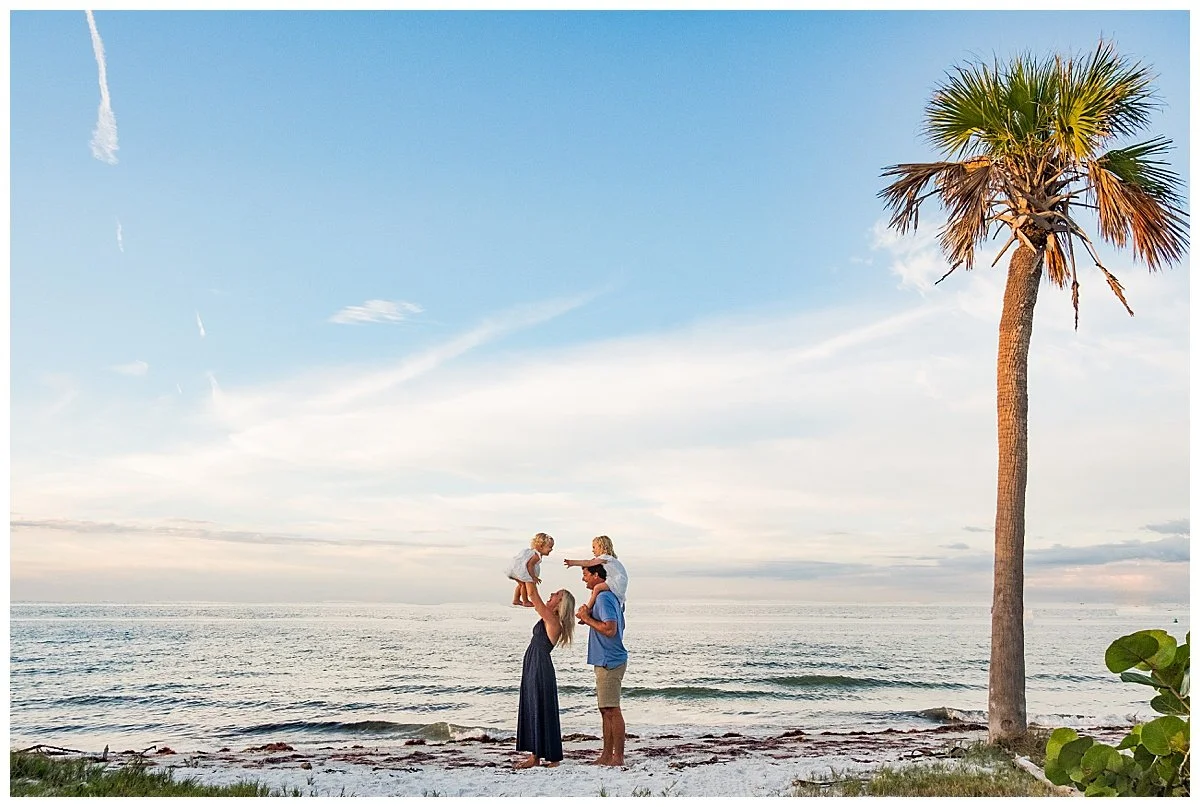 A family of four enjoying a day at the beach, with a tall palm tree nearby and the ocean in the background.