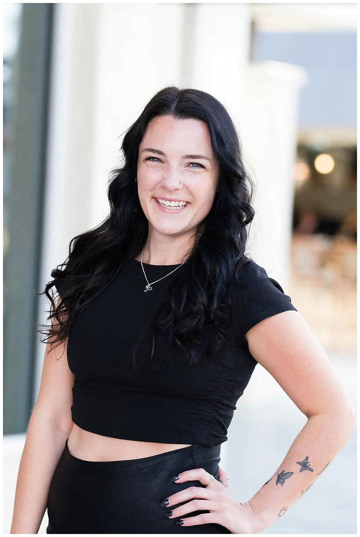 A young woman with long black wavy hair, smiling and wearing a black crop top and a black skirt, standing outdoors with her hand on her hip, showcasing butterflies tattooed on her arm.