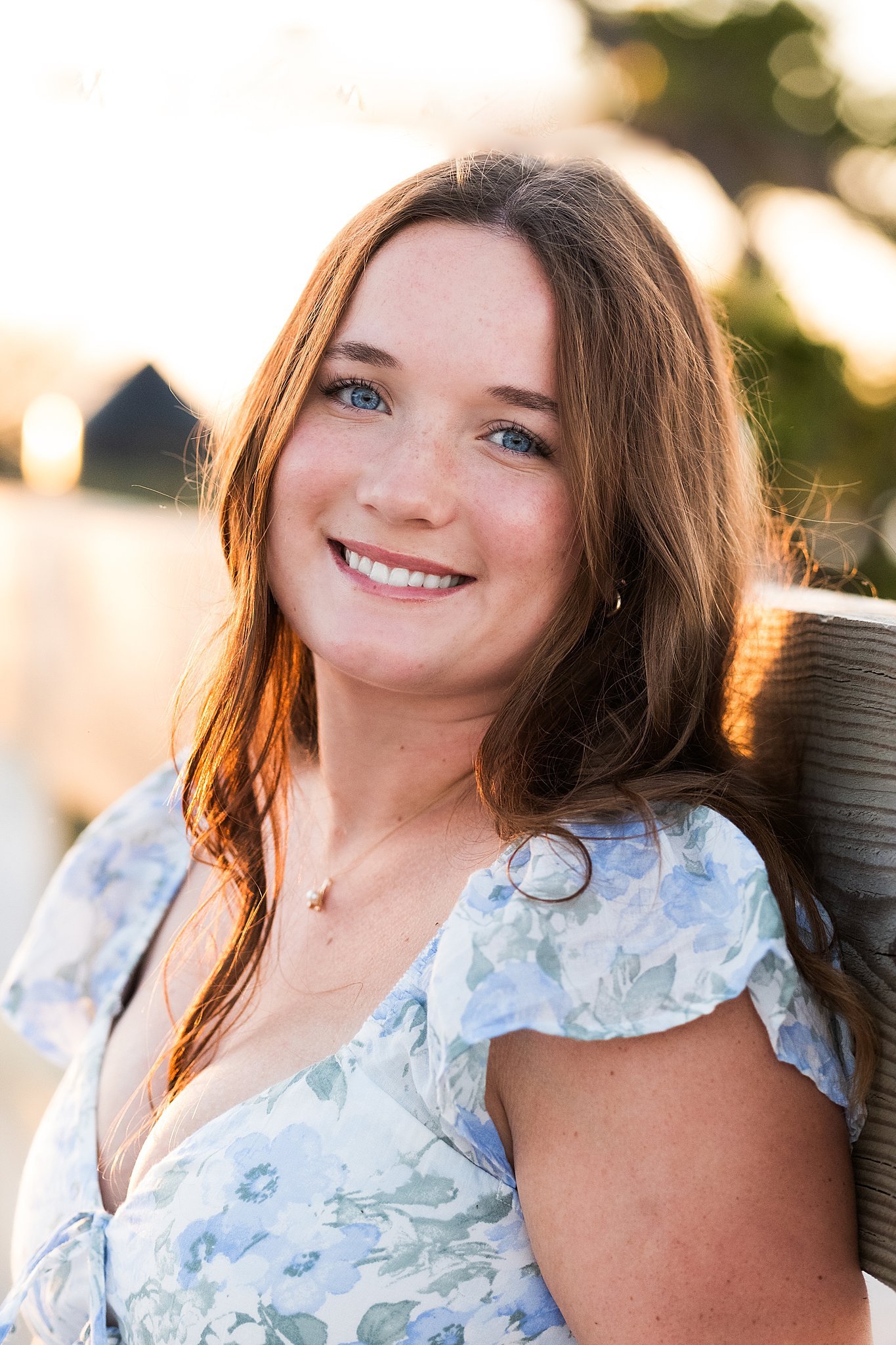 Close-up of a young woman with red hair and blue eyes smiling outdoors near a body of water at sunset, wearing a light floral dress and a necklace.