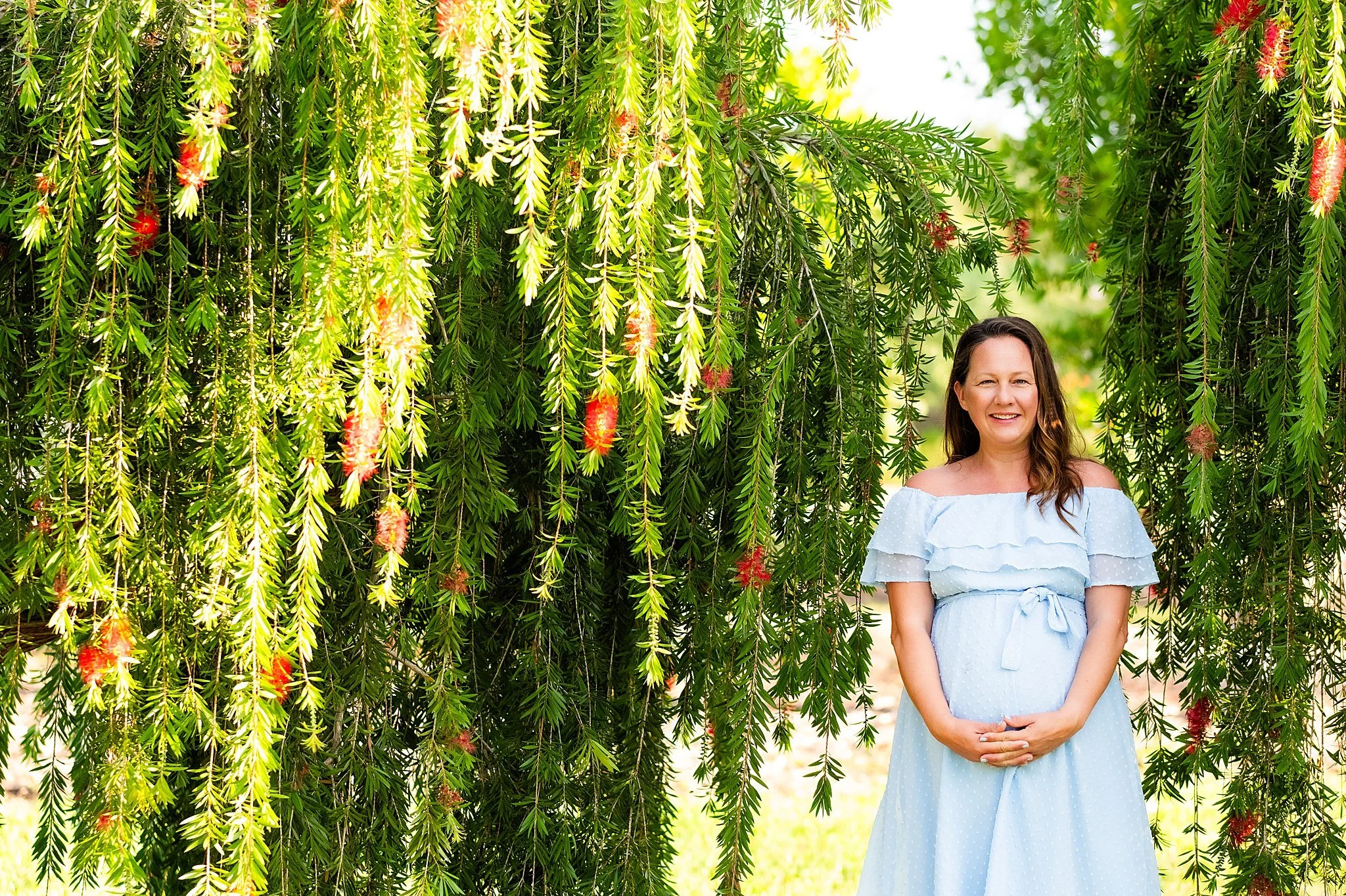 A woman wearing a light blue off-the-shoulder dress smiling, standing under hanging green wisteria vines with red flowers.