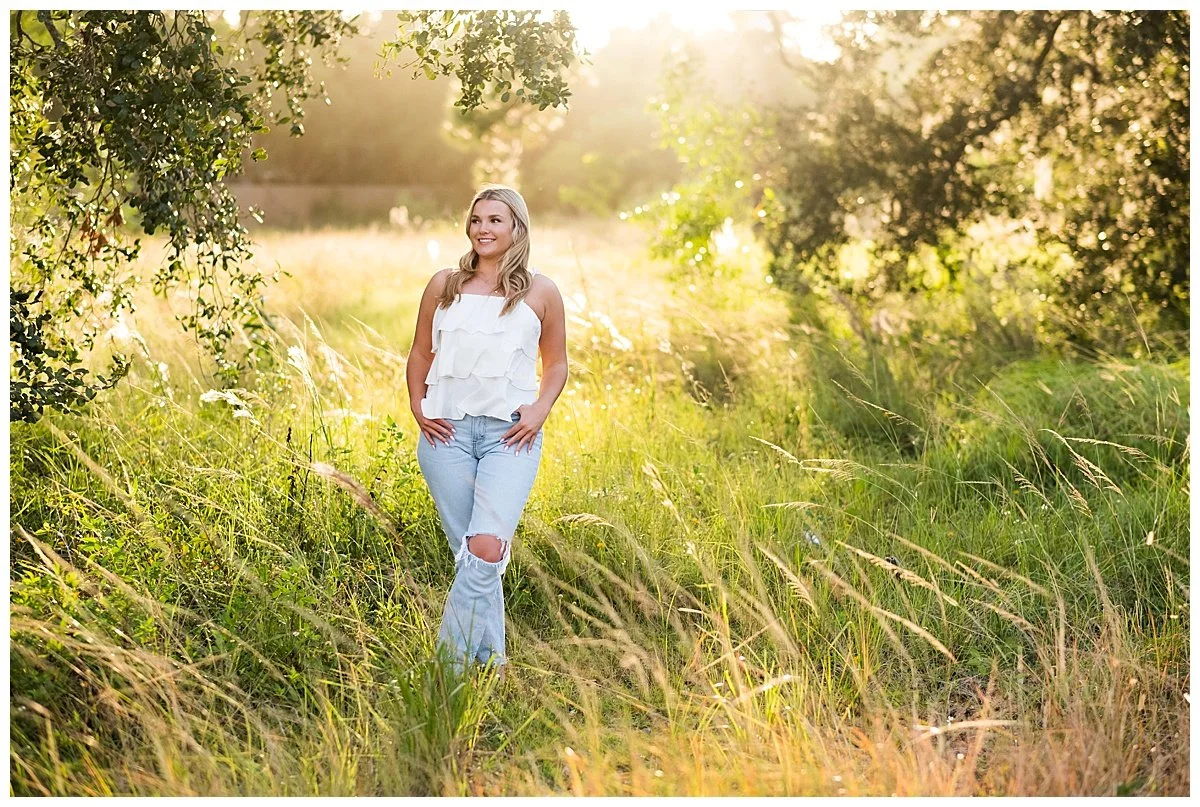 A young woman walking through a grassy field with trees on either side, dressed in a white sleeveless ruffled top and ripped jeans, smiling and enjoying the warm sunlight.