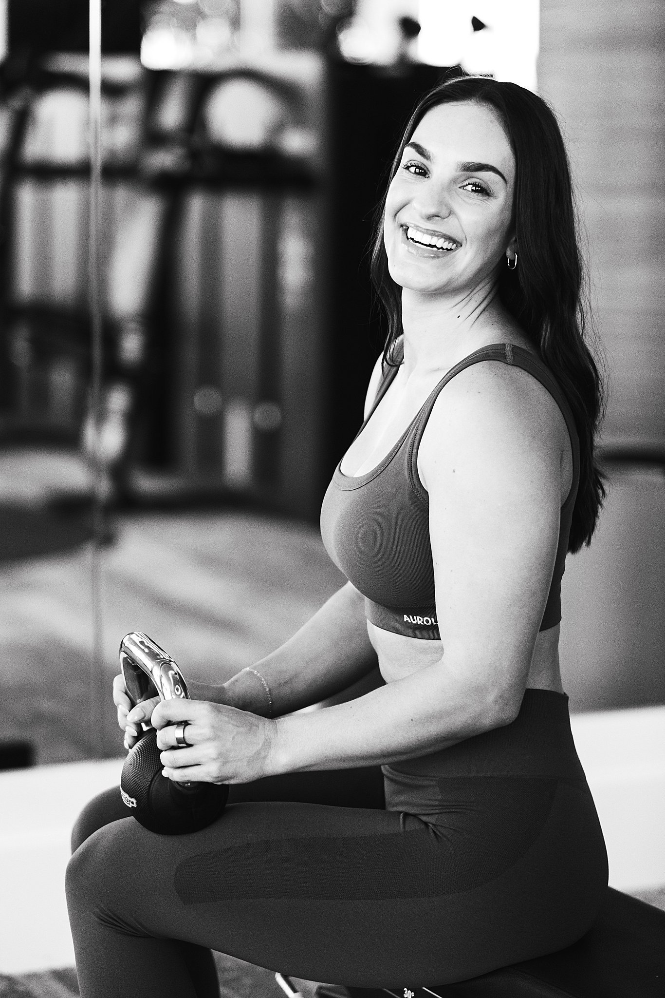 A smiling woman sitting on a workout bench holding a kettlebell in a gym.