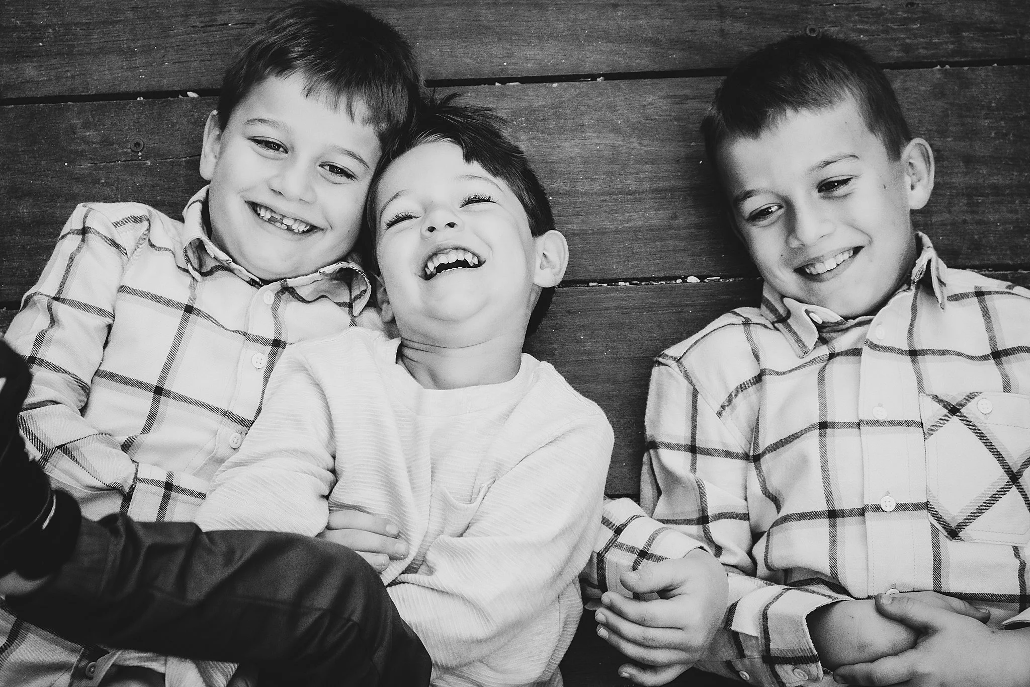 Three young boys lying on a wooden surface, smiling and laughing together.
