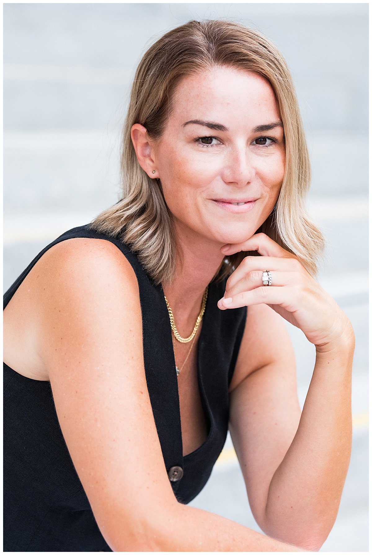 A woman with light skin and shoulder-length blonde hair, wearing a black sleeveless top, gold jewelry, and a diamond ring, smiling and posing with her hand resting on her chin against a blurred outdoor background.