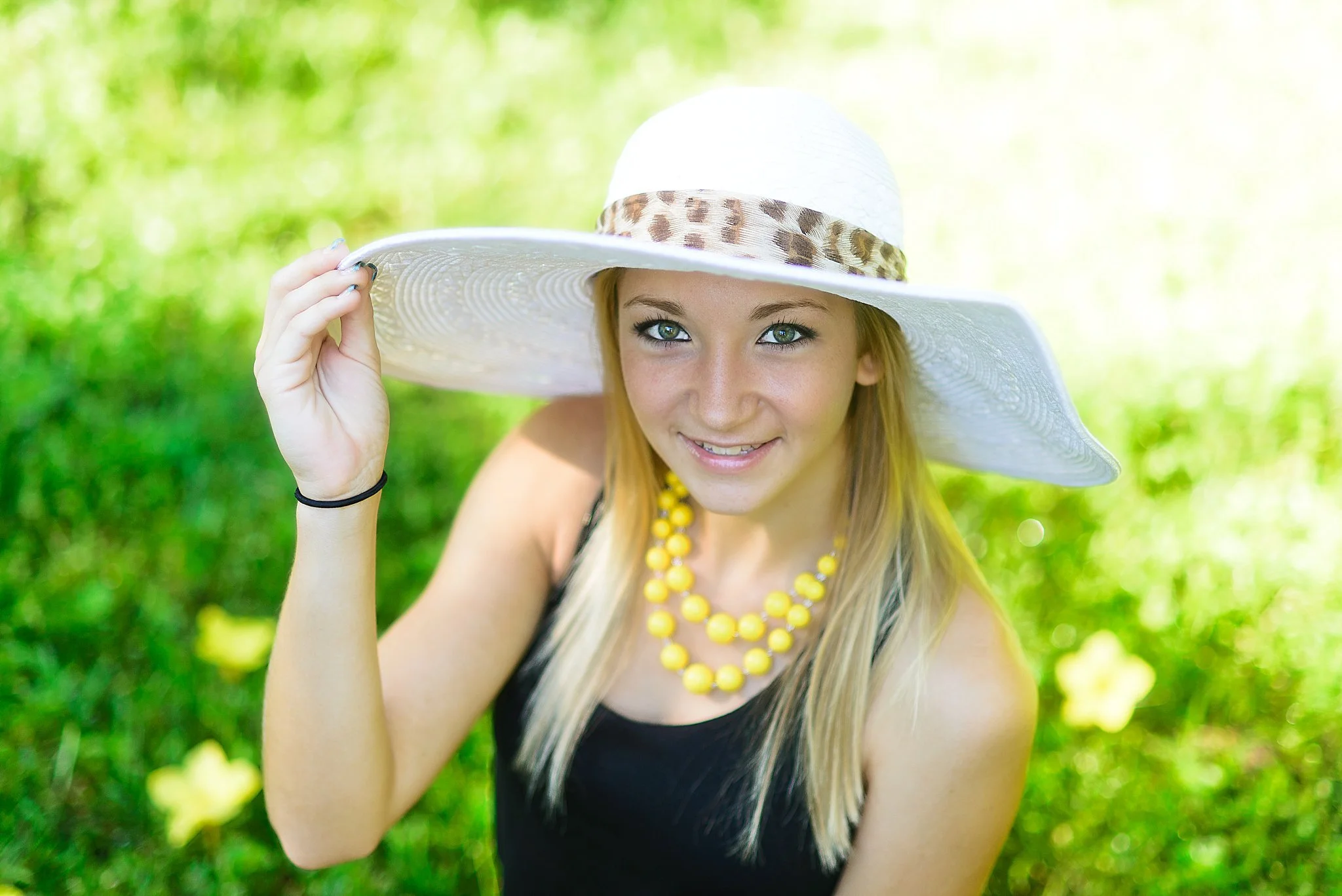 A young woman with blonde hair and blue eyes wearing a large white sun hat with a leopard print band, a black sleeveless top, and a yellow beaded necklace, smiling outdoors on a sunny day, holding the brim of her hat with her right hand, in a lush gr
