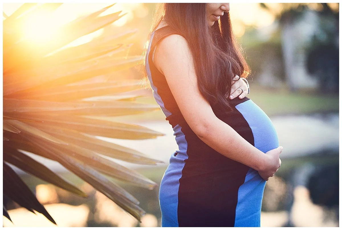 A pregnant woman stands outdoors in the sunlight, holding her belly with one hand and cradling it with the other, wearing a black and blue sports dress.