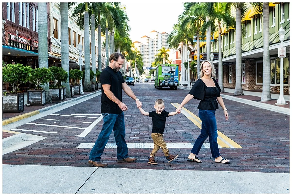 A family of three crossing a city street with palm trees, shops, and apartments in the background. The child is holding hands with his parents and smiling, while the father and mother hold the child's other hands and walk on either side.