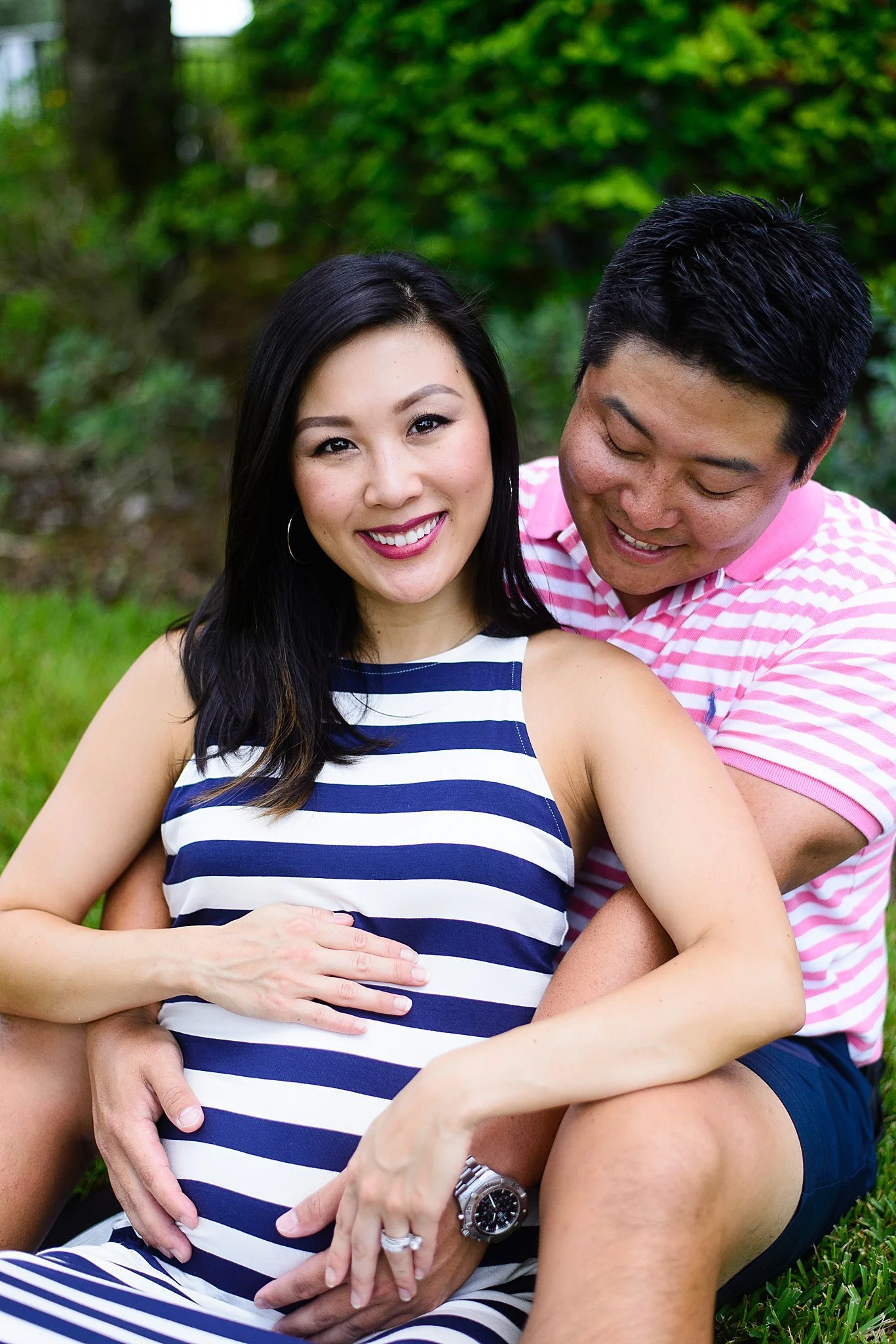 A happy pregnant woman with long black hair, wearing a navy and white striped sleeveless dress, sitting on grass outdoors with a man behind her, both smiling. The man, with short black hair, wearing a pink and white striped polo shirt, embraces her f