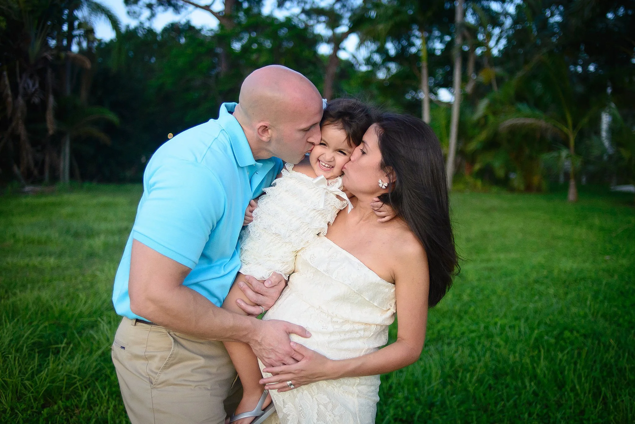 A family of three, a man, woman, and young girl, sharing a loving moment outdoors during daytime, with the man and woman giving kisses to the girl, all smiling.