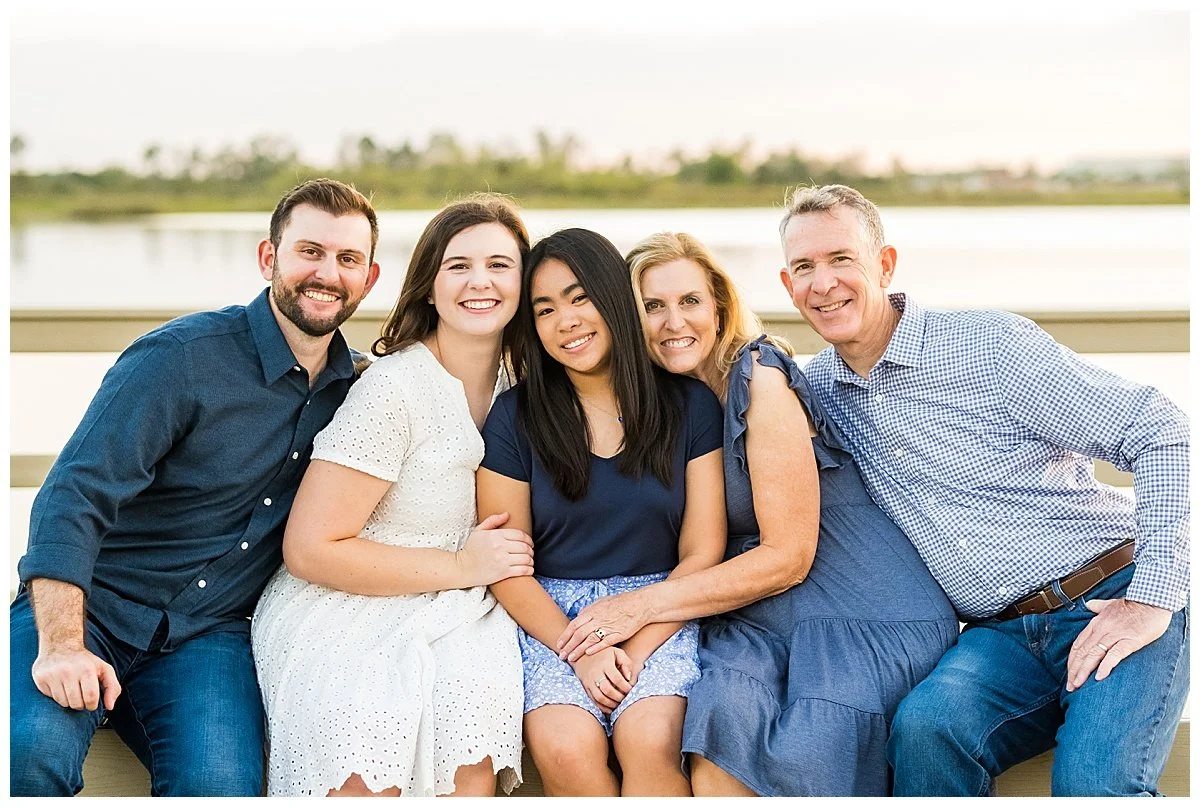 Family of five sitting on a bench outdoors by a lake during sunset, smiling at the camera, dressed in casual summer clothing.