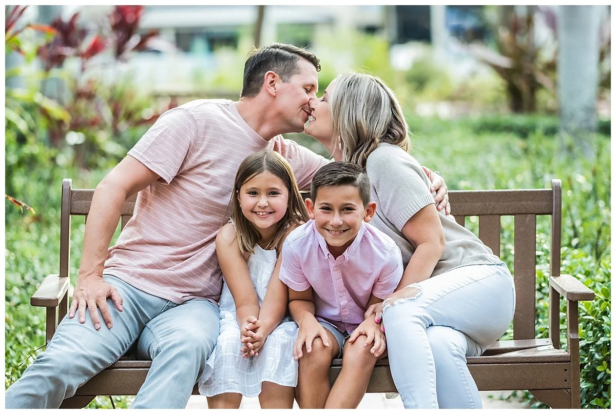 A family of four sitting on a wooden park bench, with the father and mother leaning in for a kiss, while the children, a girl and a boy, sit between them smiling.