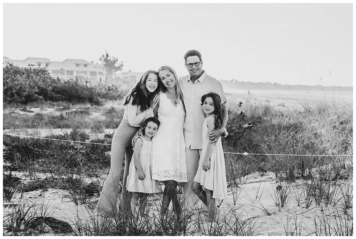 A family of six standing on a sandy beach with grass, smiling and hugging, in front of a background of buildings and open sky.
