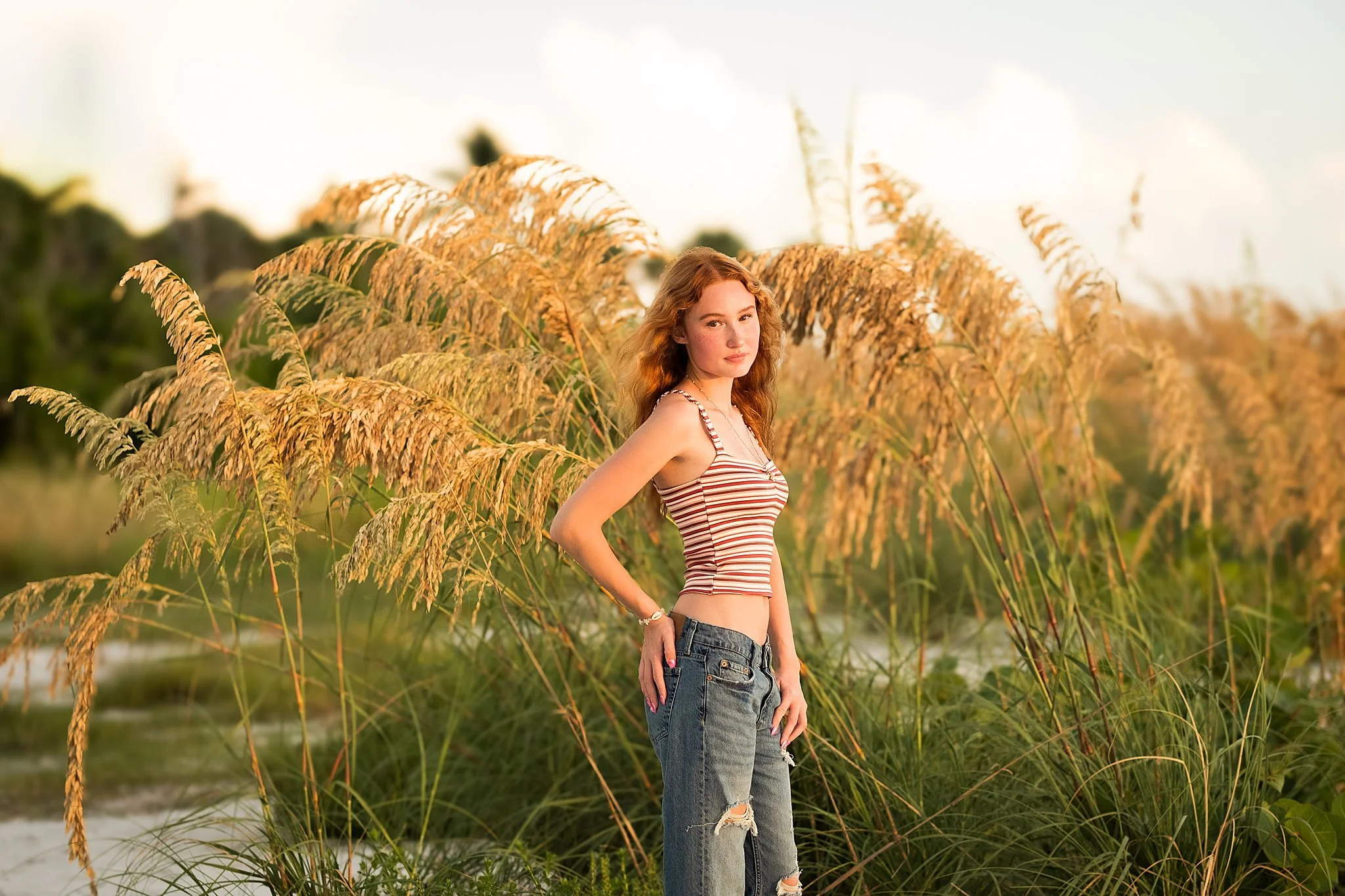 Young woman with red hair in a striped tank top and ripped jeans standing among tall grasses at sunset.