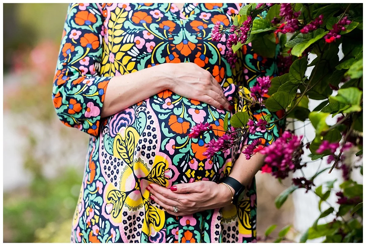 Close-up of a pregnant woman's belly and hands outdoors with pink flowers and greenery, wearing a colorful floral dress.