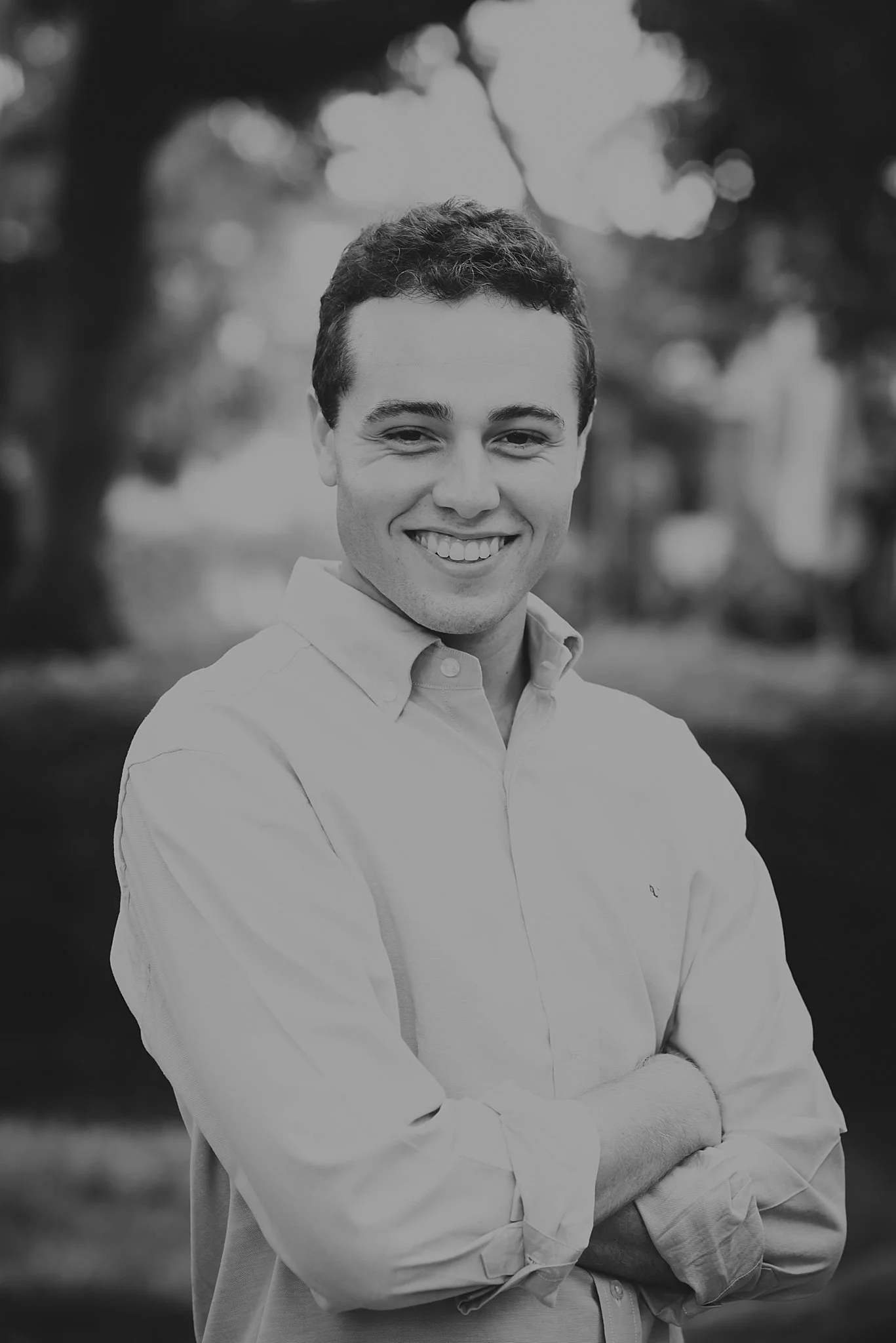 Black and white portrait of a young man smiling with arms crossed, outdoors with trees in the background.
